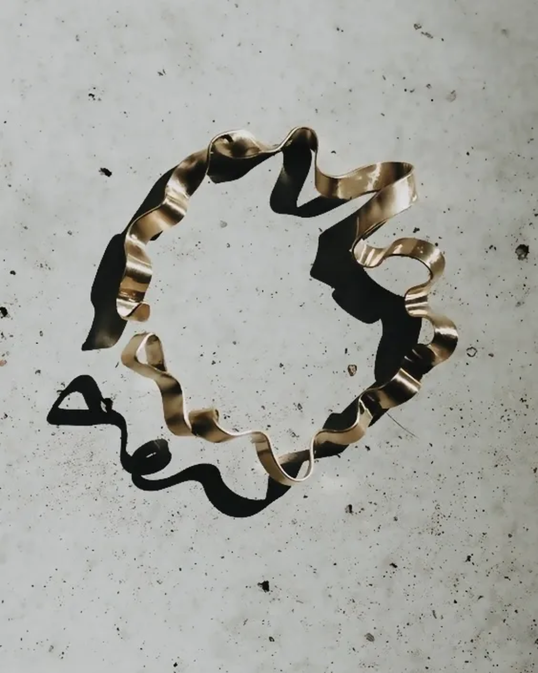 A metallic gold wavy ring-shaped hair claw clip or scrunchie-like accessory laid flat on a light gray textured surface, casting a dark shadow.
