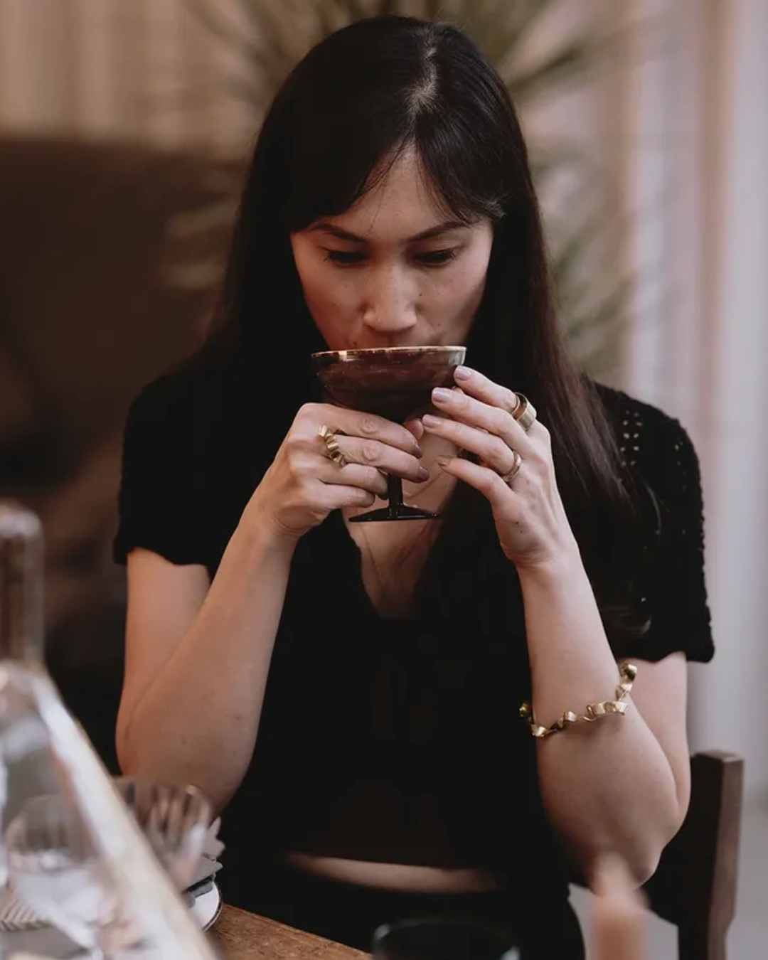 A woman in a black short-sleeve top holding a dark stemmed glass, wearing gold rings and a gold beaded bracelet, in a warmly lit indoor setting.