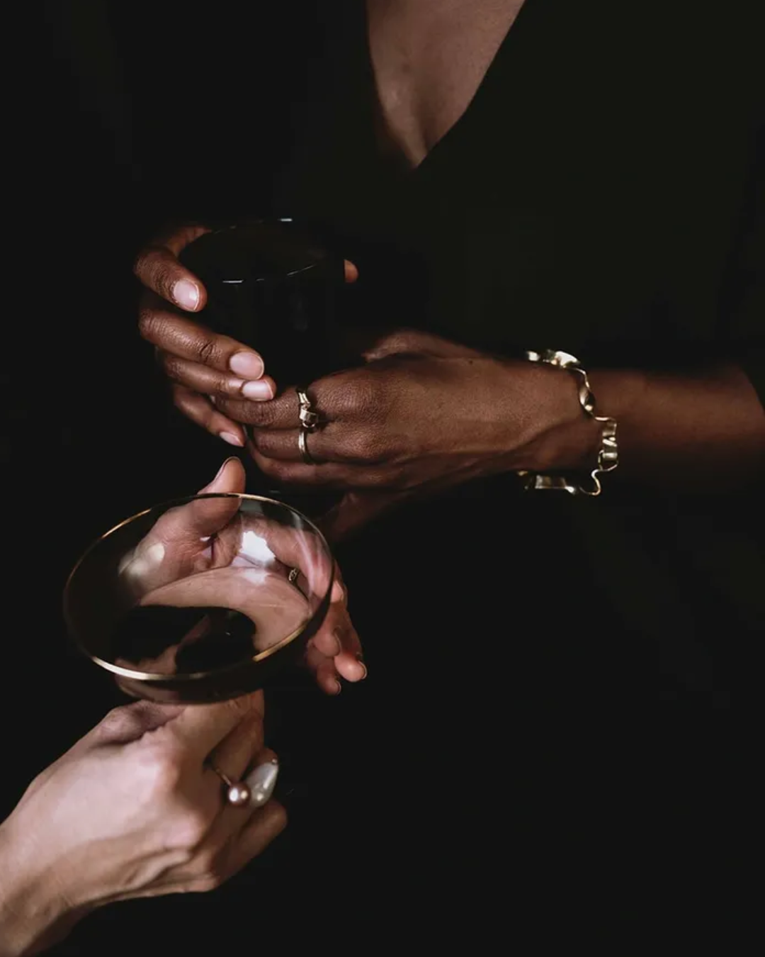 Close-up of hands holding a black tumbler and a gold-rimmed coupe glass against a dark background, with rings and bracelets visible on the fingers.
