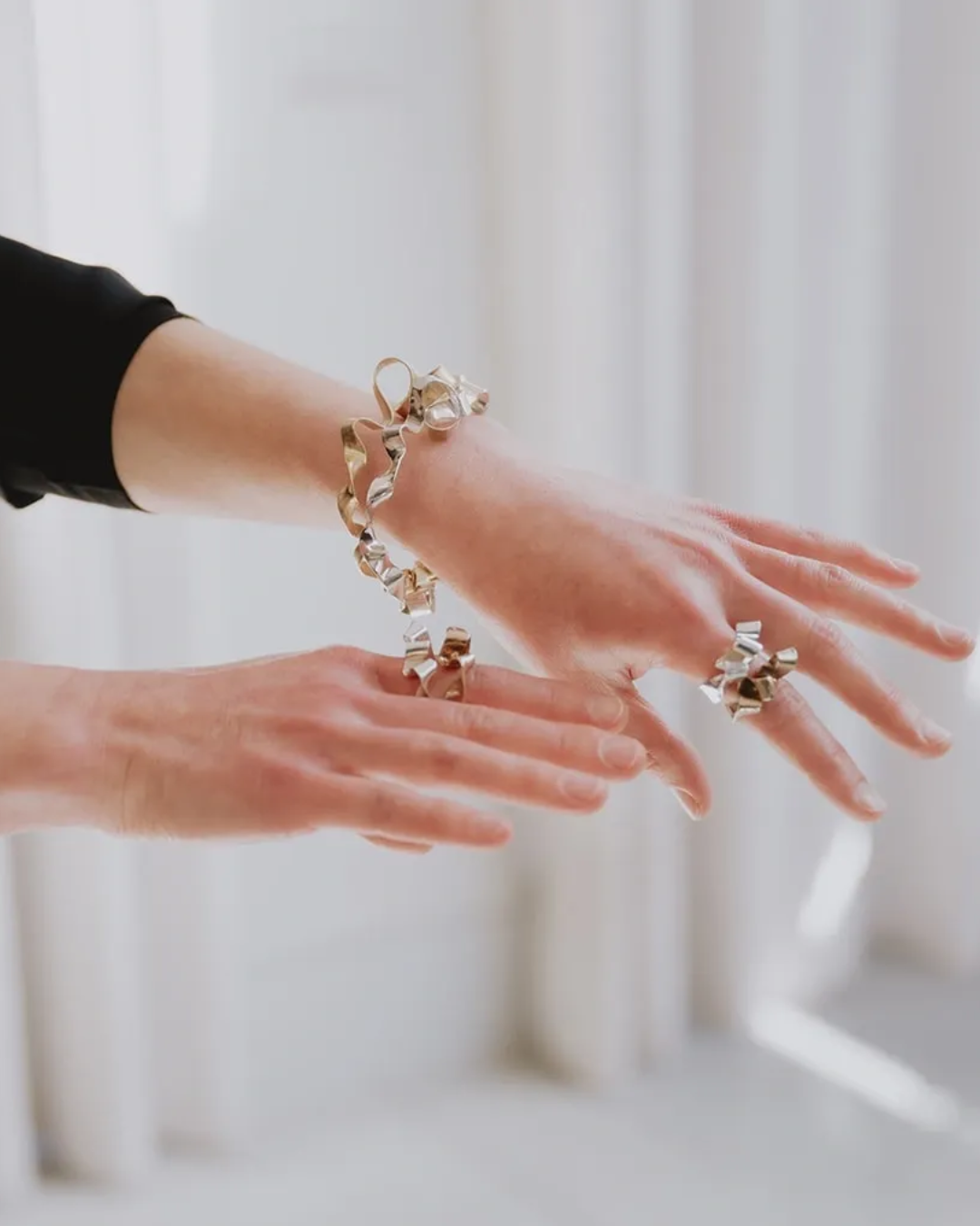 A close-up lifestyle shot of a person's hands and wrists wearing a matching gold jewelry set featuring a bracelet and rings with sculptural, flower-like metallic details.