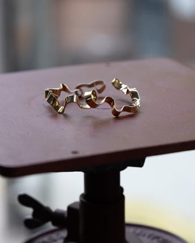 A gold-toned, wavy open cuff ring displayed on a dark brown wooden stand against a softly blurred background.