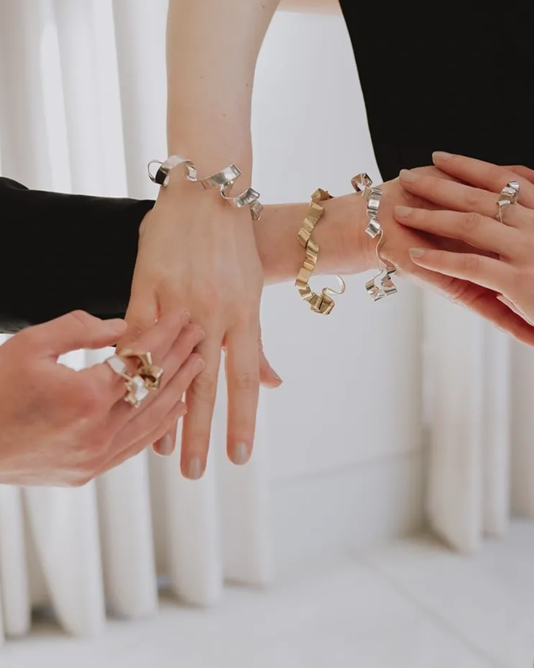 Close-up of multiple hands wearing and adjusting chunky geometric jewelry, including silver and gold twisted link bracelets and rings, against a soft white curtain background.