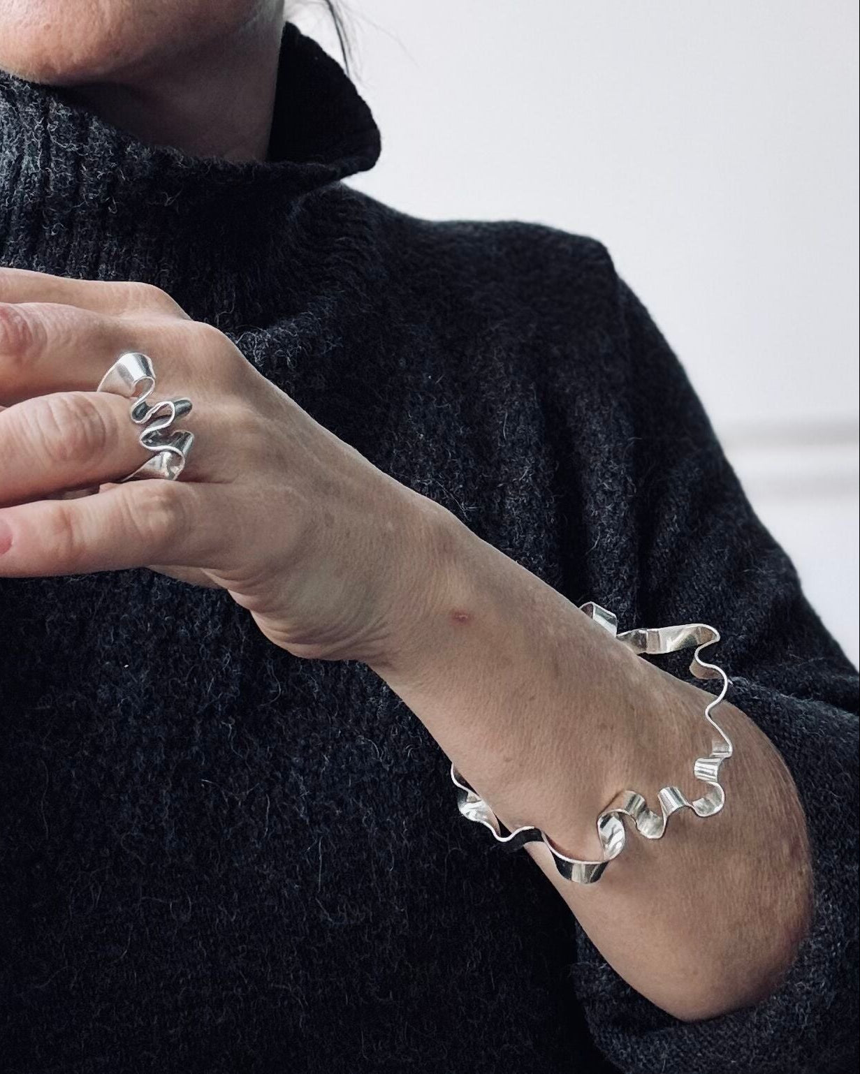 A close-up of a person wearing a matching silver ring and bracelet with an abstract, wavy organic design, paired with a dark charcoal knit turtleneck sweater.