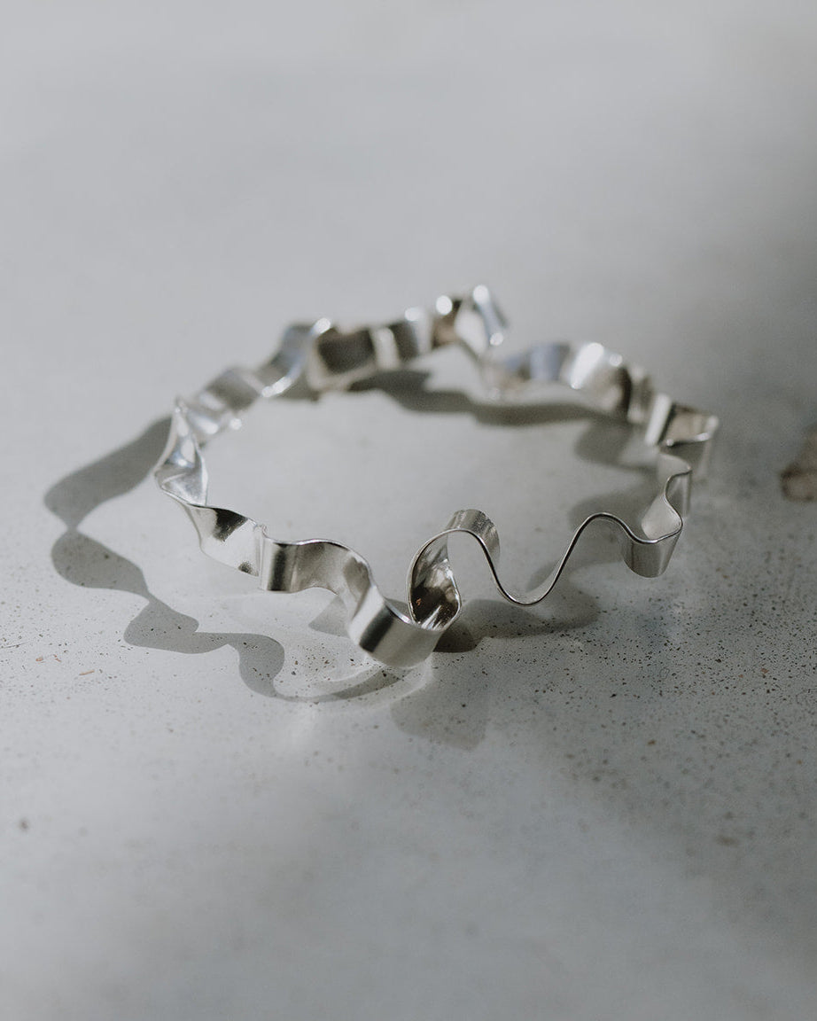 A polished silver wavy bangle bracelet resting on a textured light gray surface, casting a soft shadow.