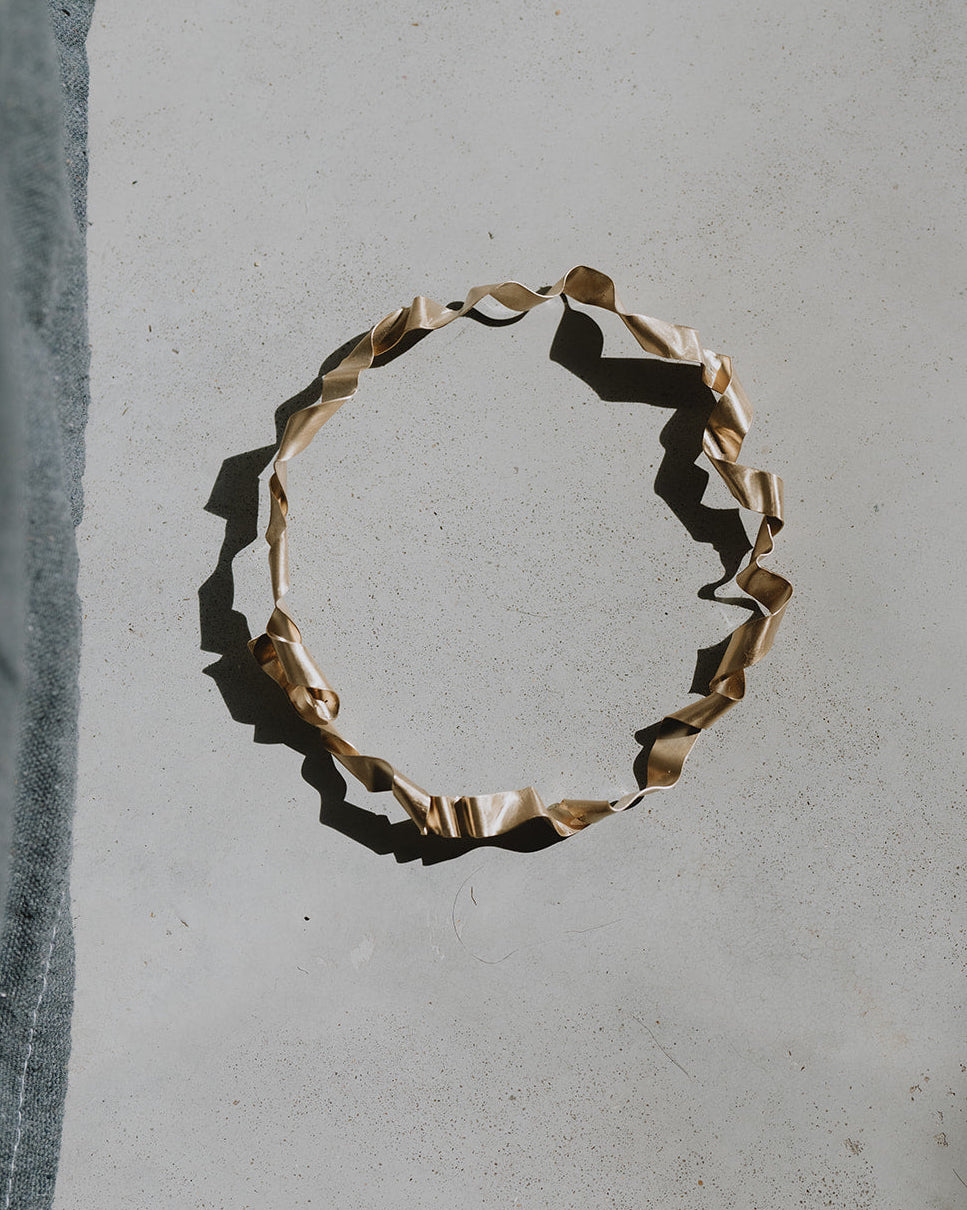 A twisted, organic-shaped gold ring, likely a paperclip or ribbon-wrapped accessory, displayed on a textured gray concrete surface with a strong diagonal shadow.
