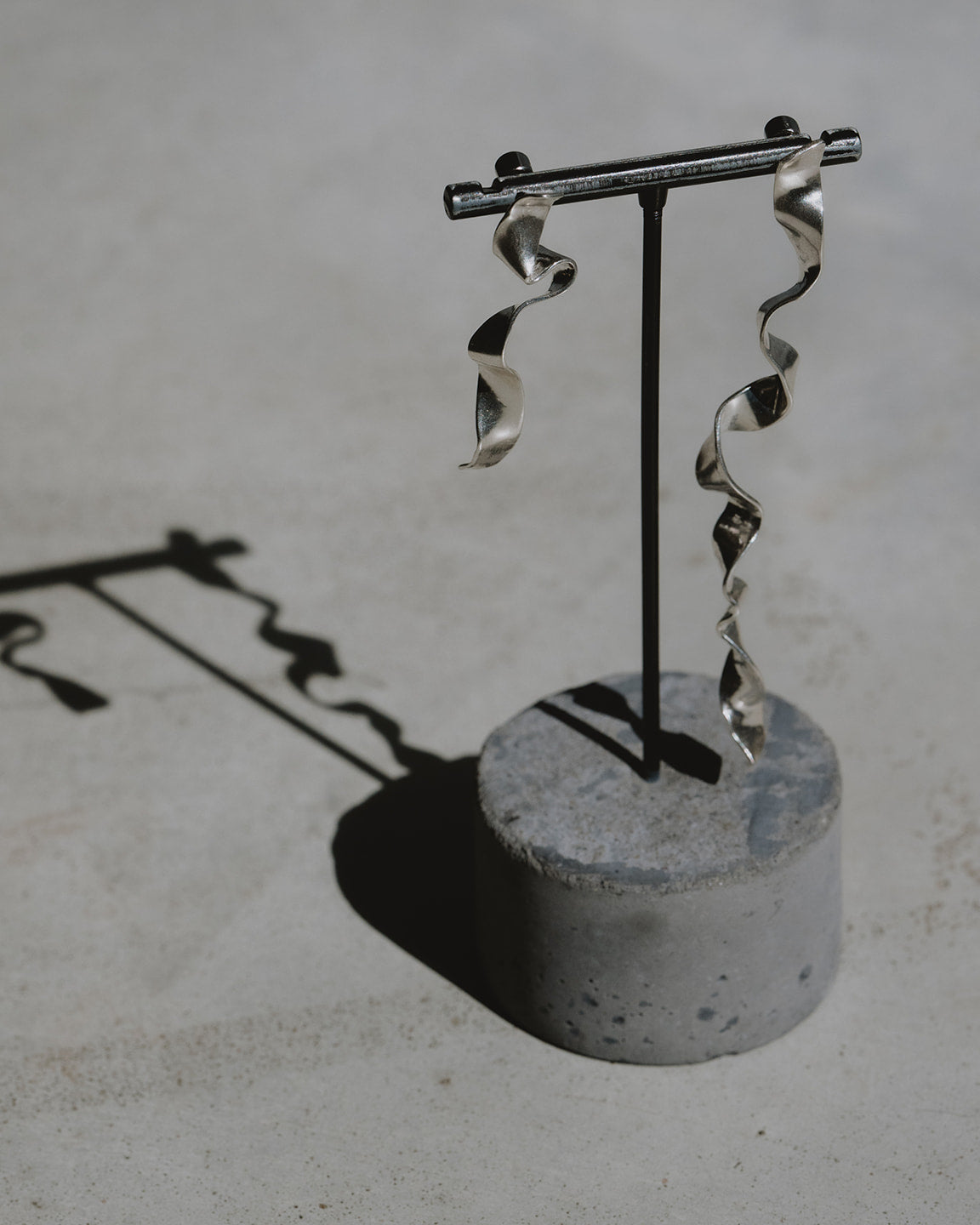 A pair of silver spiral dangle earrings displayed on a black stand with a round concrete base, photographed on a light gray textured surface with a sharp shadow.
