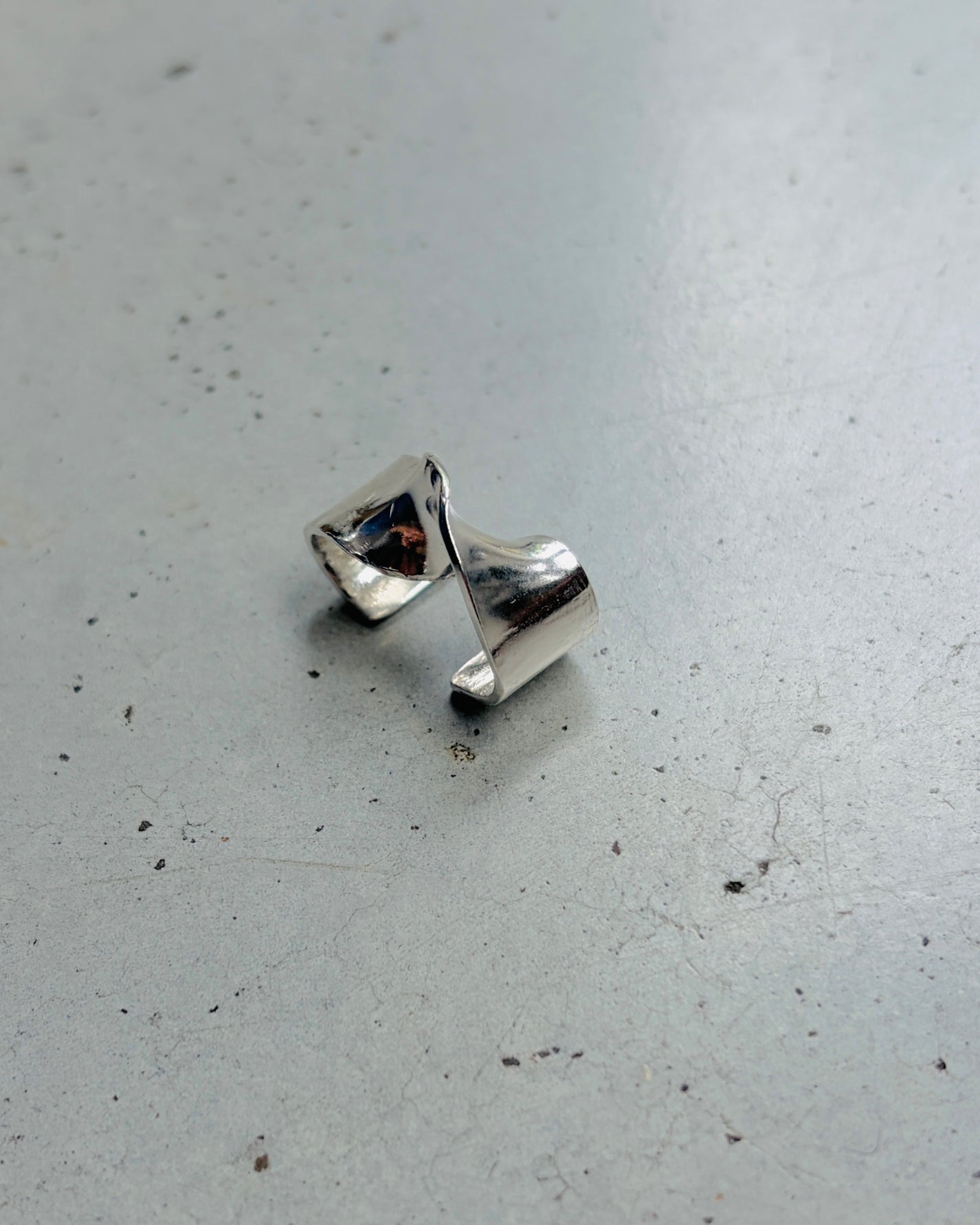 A polished silver ring with a sculptural, twisted triangular design, resting on a textured gray concrete surface.