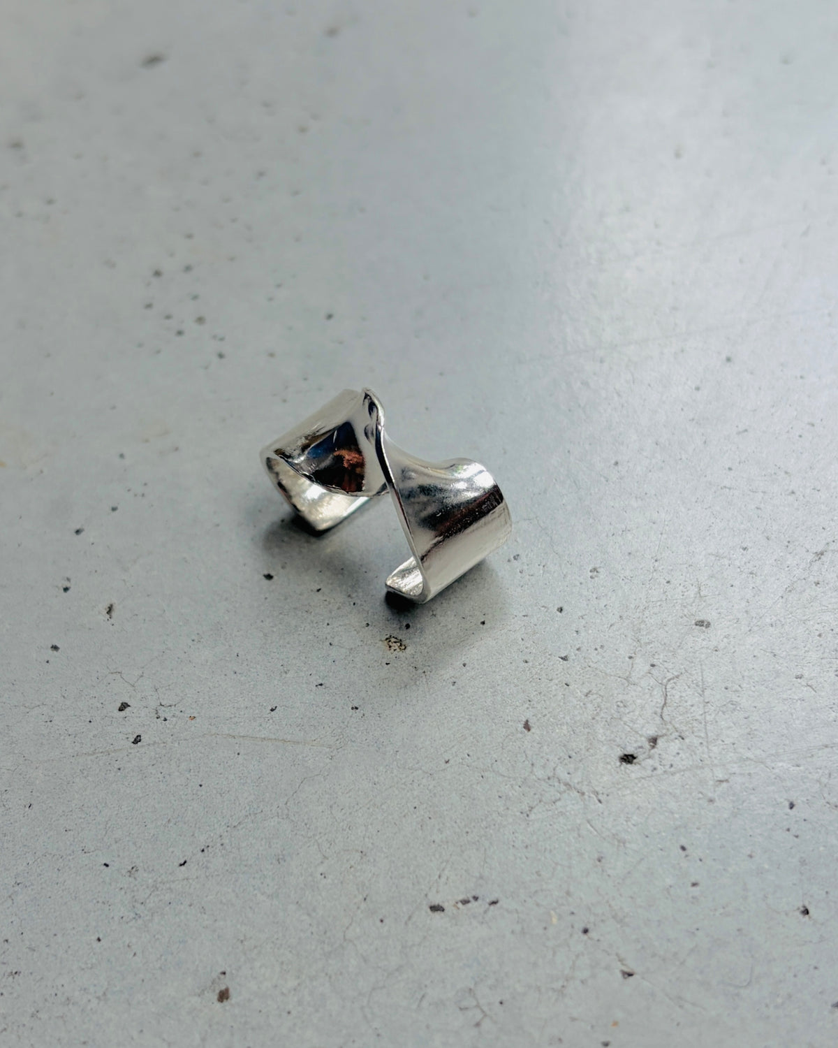 A polished silver ring with a sculptural, twisted triangular design, resting on a textured gray concrete surface.
