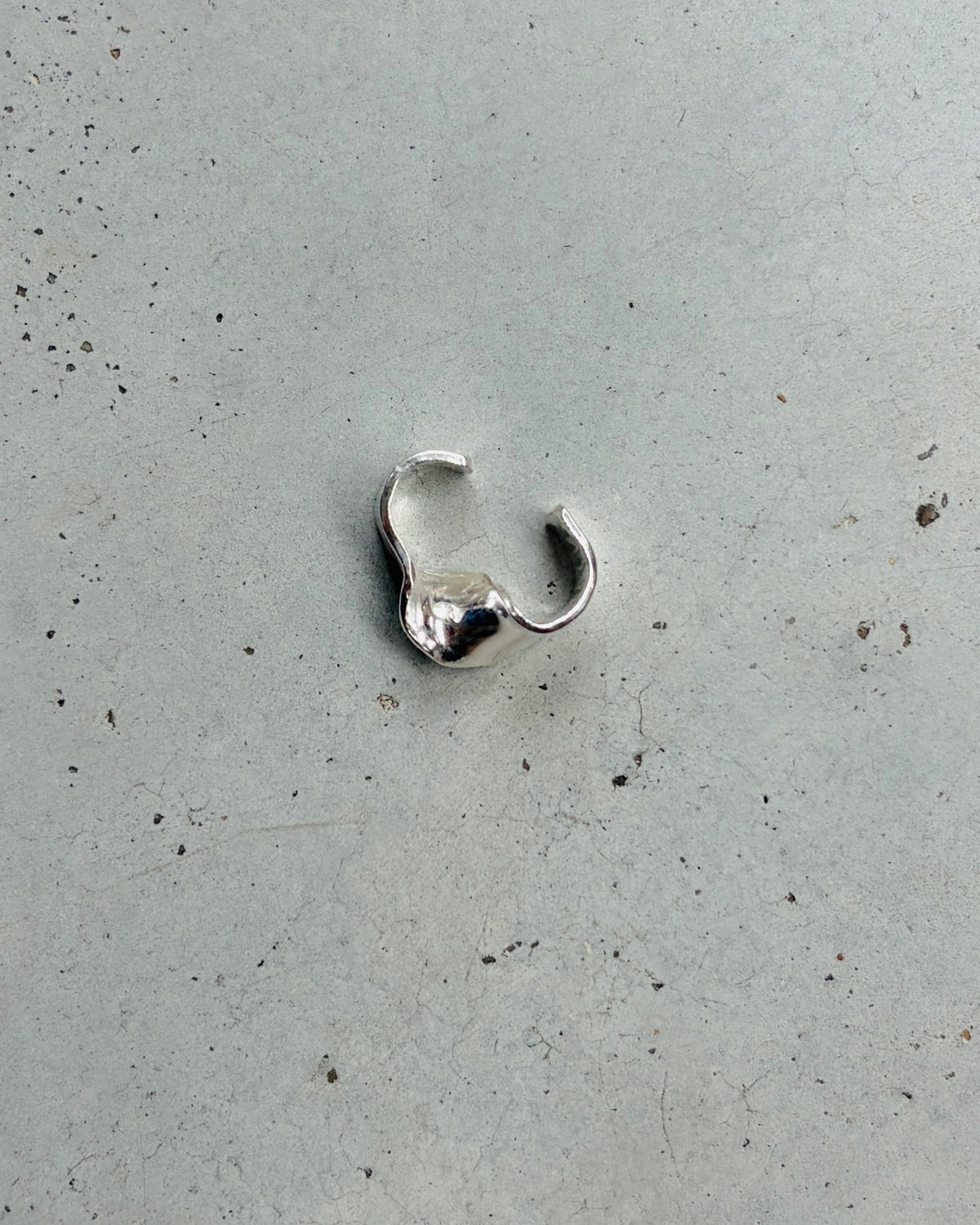 A small silver-toned metal ring with a textured, organic wavy shape, placed on a light gray concrete surface.