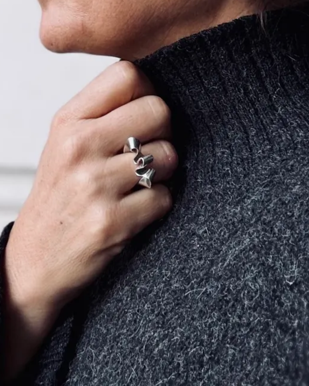 A close-up of a person wearing a chunky silver ring with three twisted, spiral-like shapes, styled with a dark gray ribbed knit sweater.