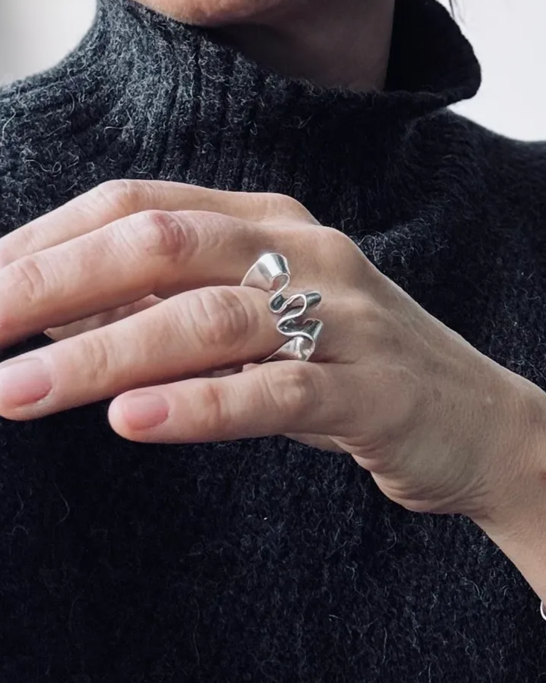 A close-up of a person's hand wearing a silver ring with a wavy, layered design, set against a dark textured knit sweater.