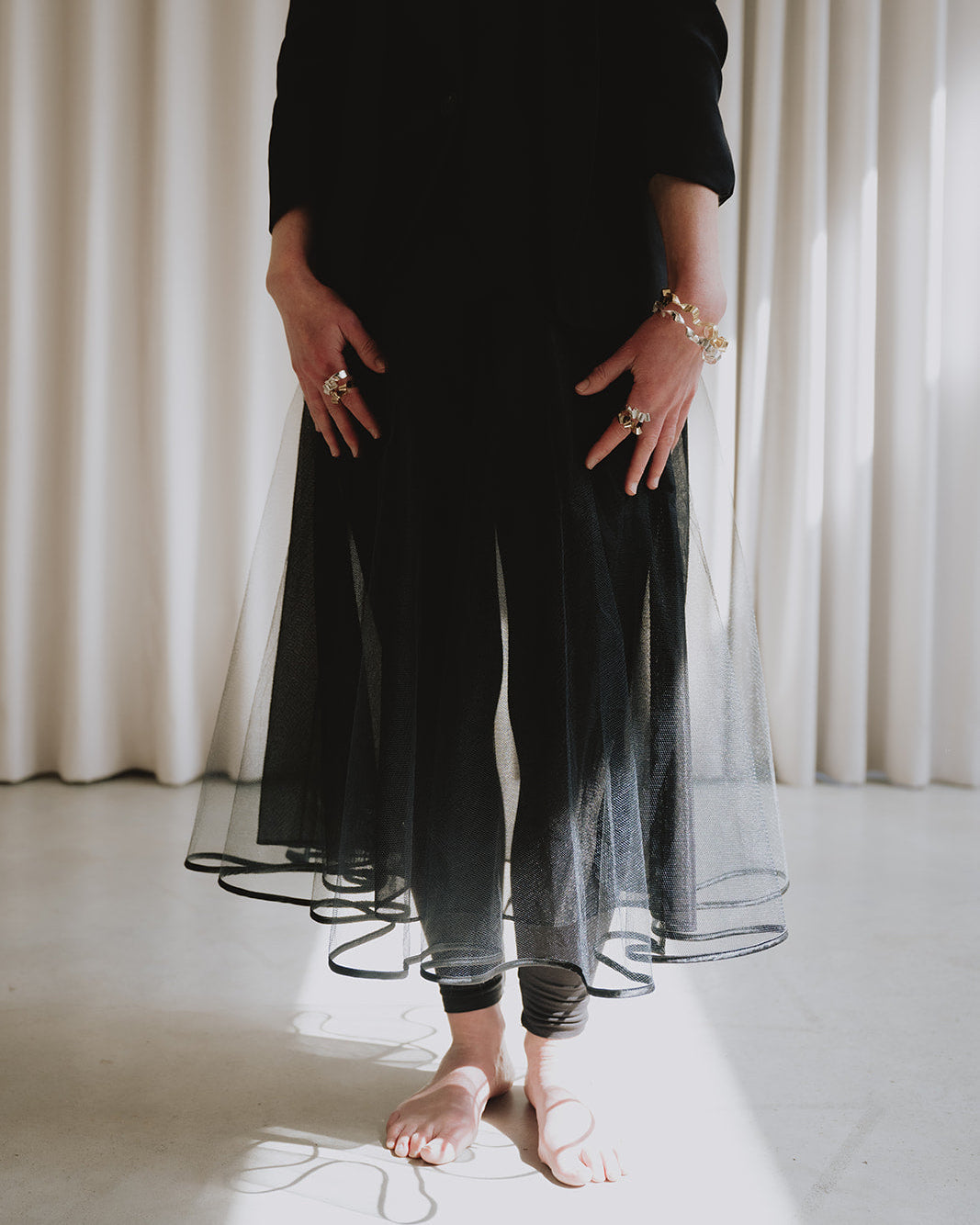 A person wearing a long black sheer tulle dress or tunic with black leggings, standing barefoot in a bright room with white curtains and sunlight on the floor.