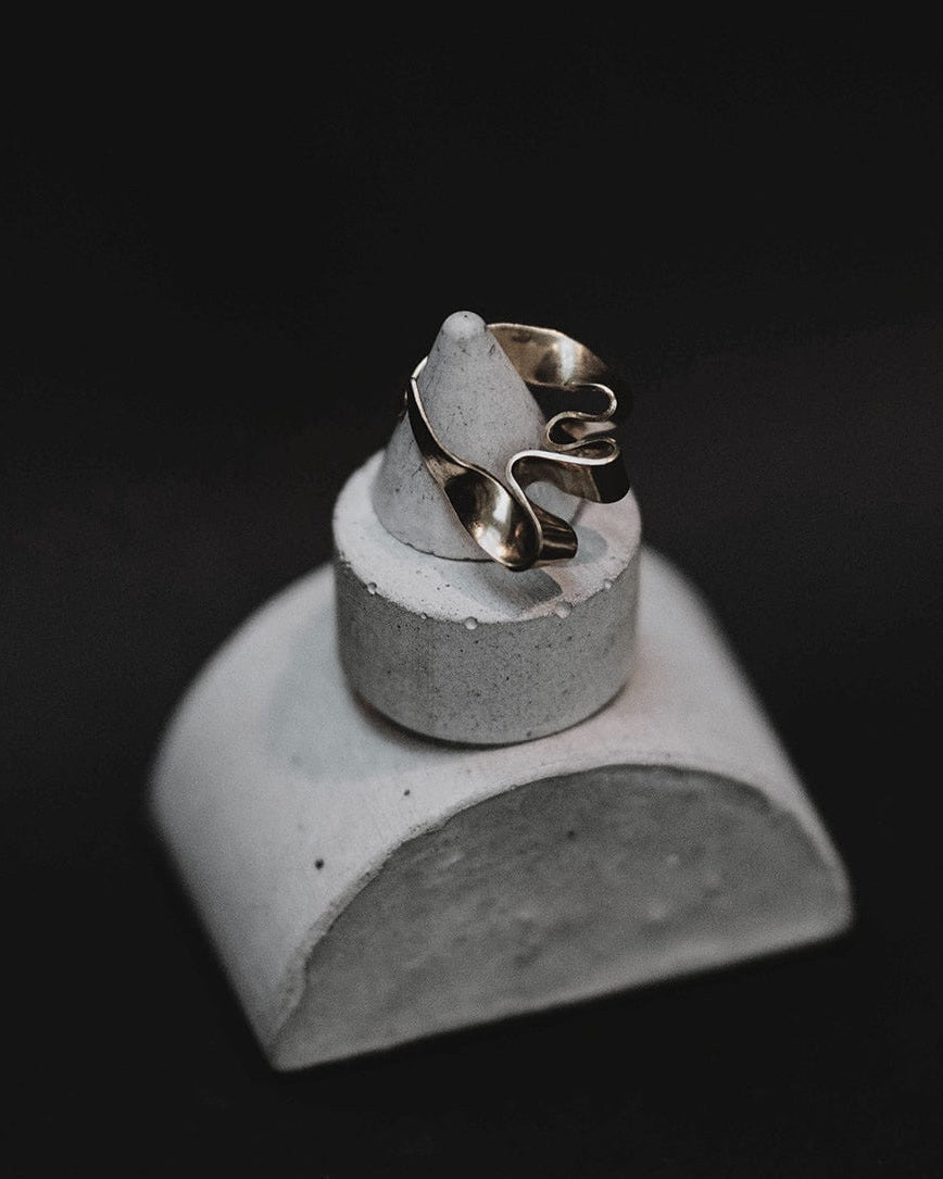 A silver-toned ring with an organic, flowing sculptural design, displayed on a small gray concrete stand against a solid black background.