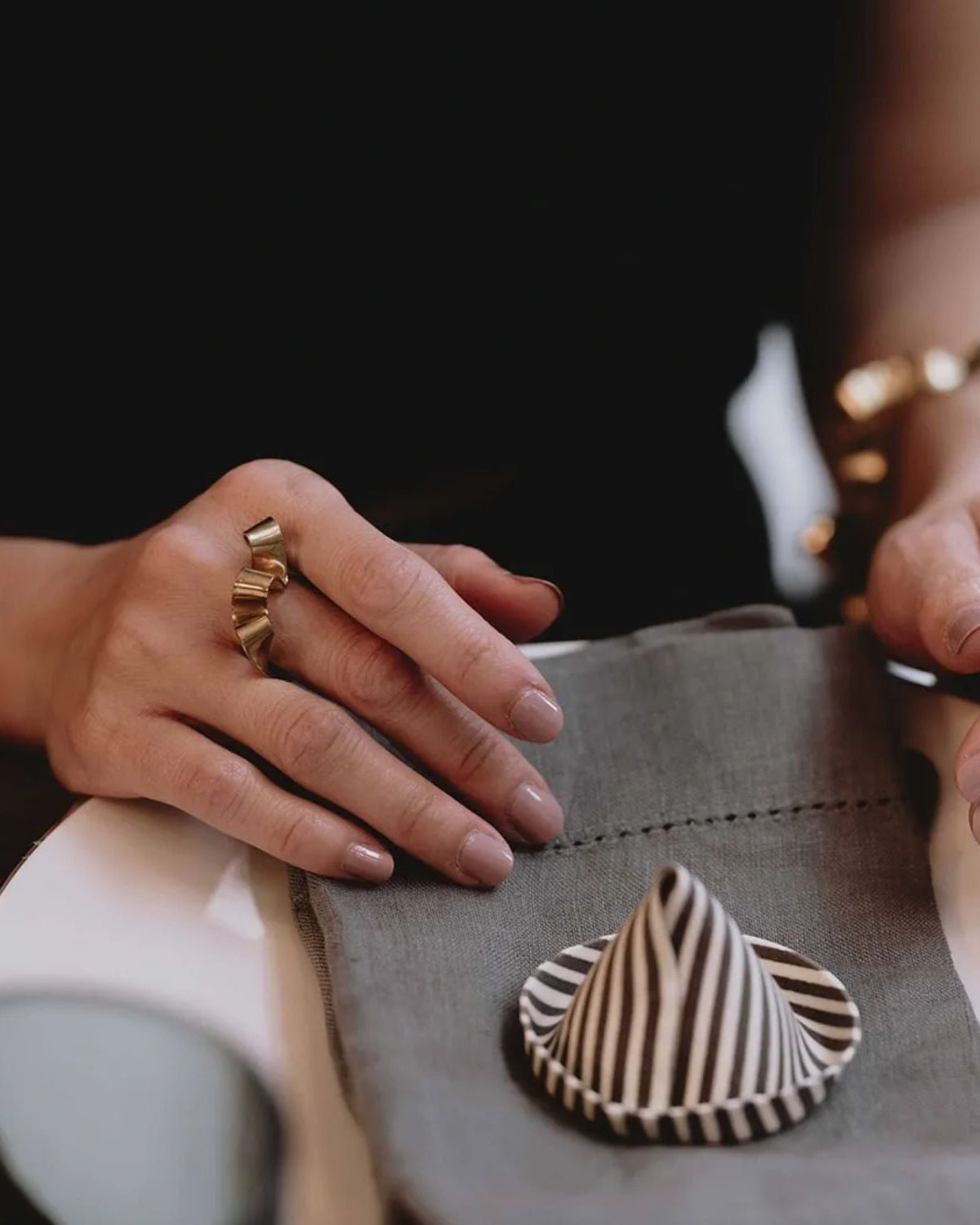 A close-up lifestyle shot of a person's hands resting on a gray fabric pouch, featuring a small striped cone-shaped accessory on top. The person is wearing multiple gold rings and bracelets.