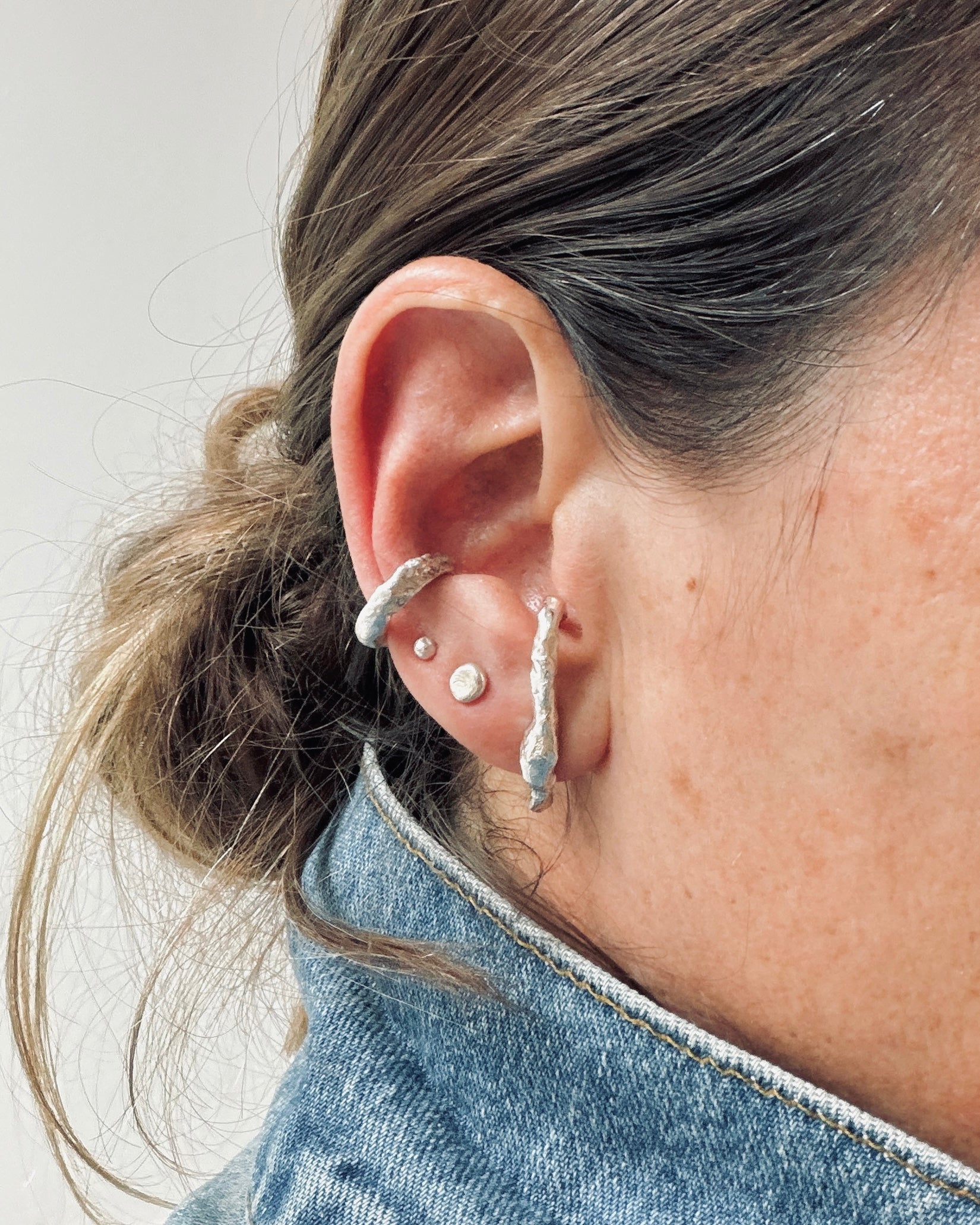 A close-up of a person's ear showcasing multiple silver earrings, including textured hoop and stud earrings, paired with a denim jacket.