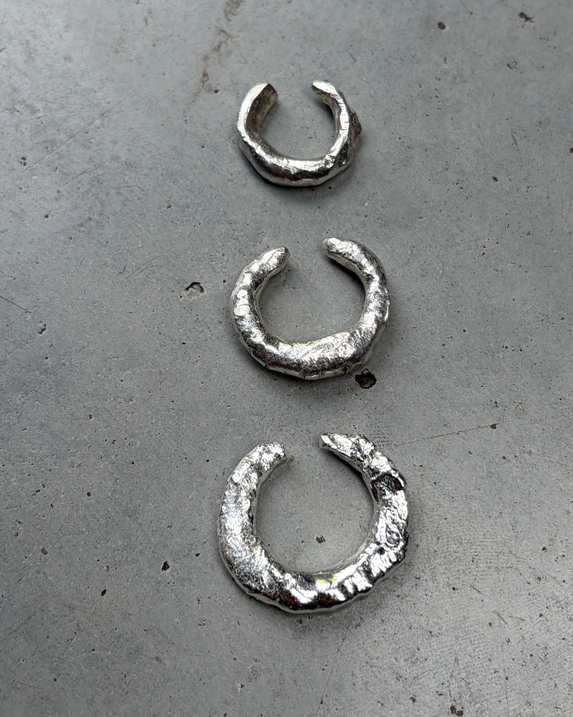 Three textured silver huggie hoop earrings in small, medium, and large sizes, arranged horizontally on a gray concrete surface.