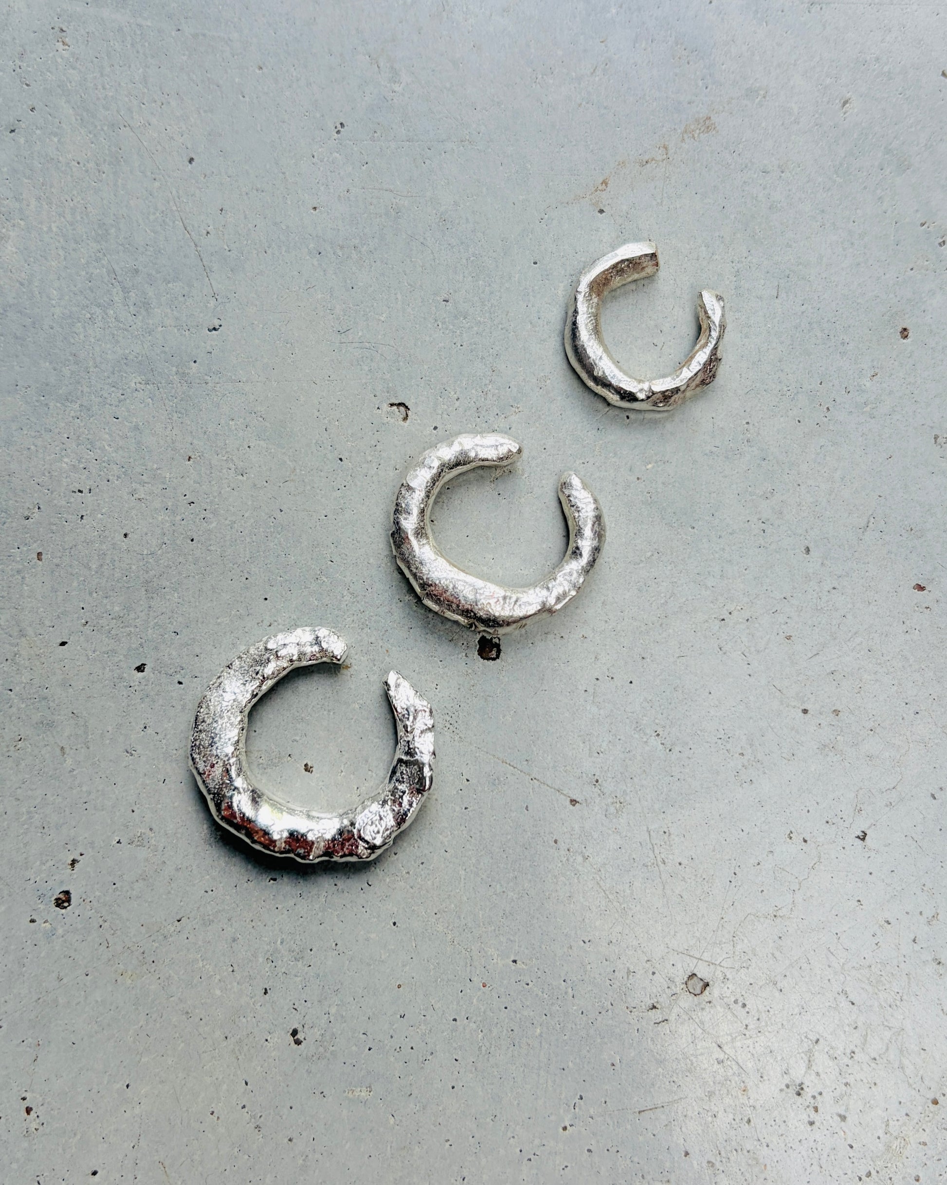 Three textured silver crescent-shaped earrings arranged diagonally on a gray concrete surface.