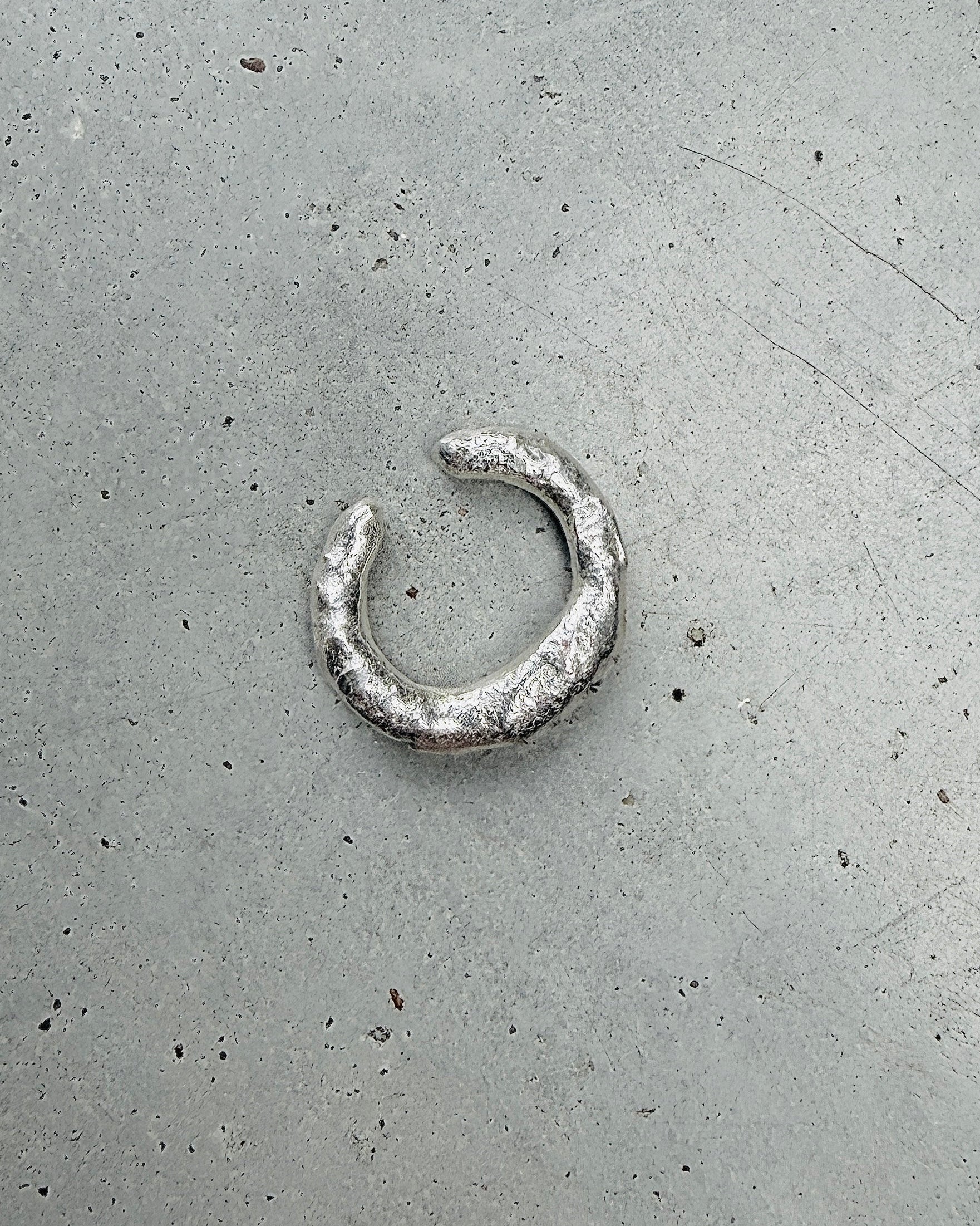 A textured silver crescent-shaped ring with a rustic, hammered finish, displayed on a speckled gray concrete surface.