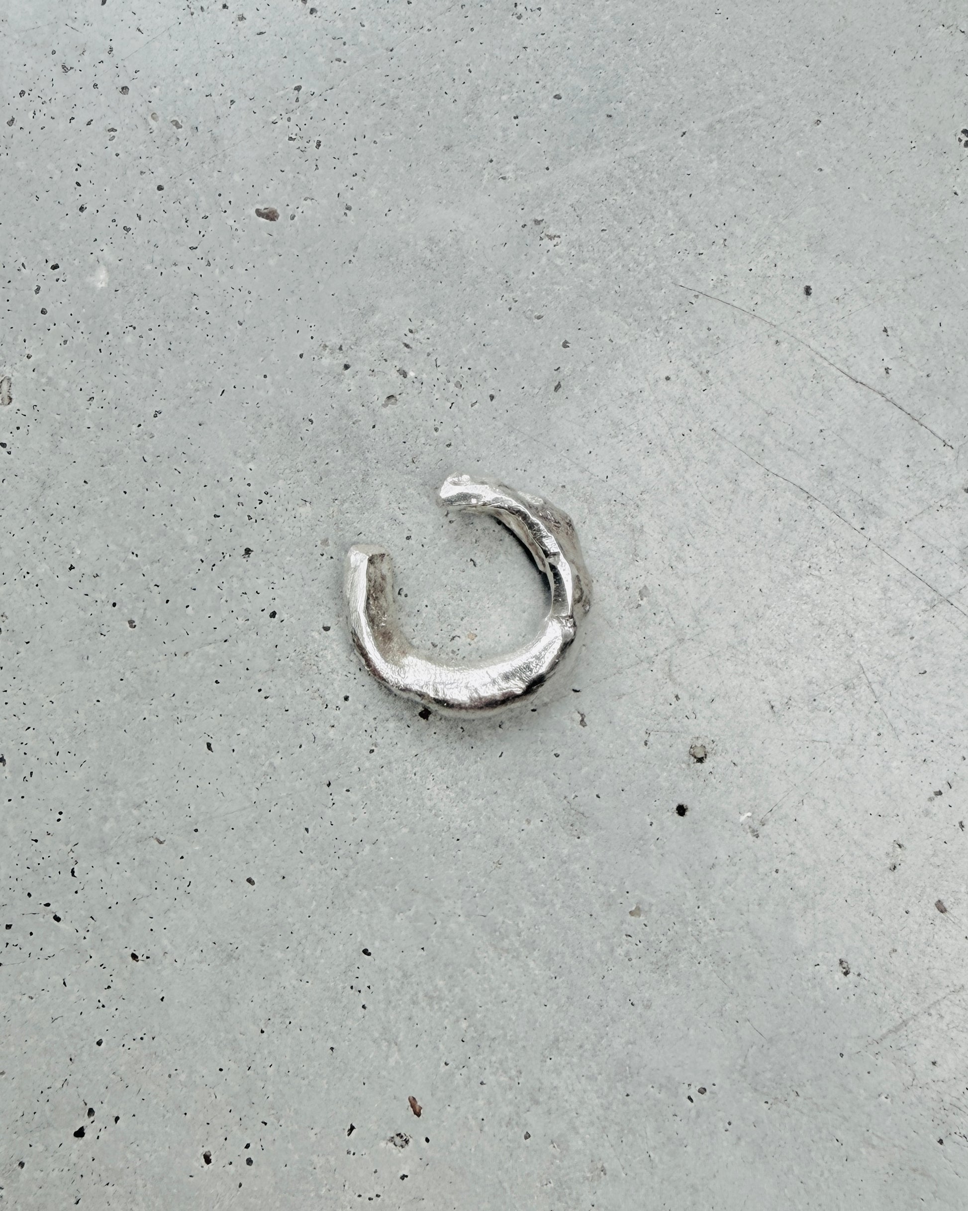 A small, textured silver crescent-shaped ring or pendant lies on a speckled gray concrete surface.
