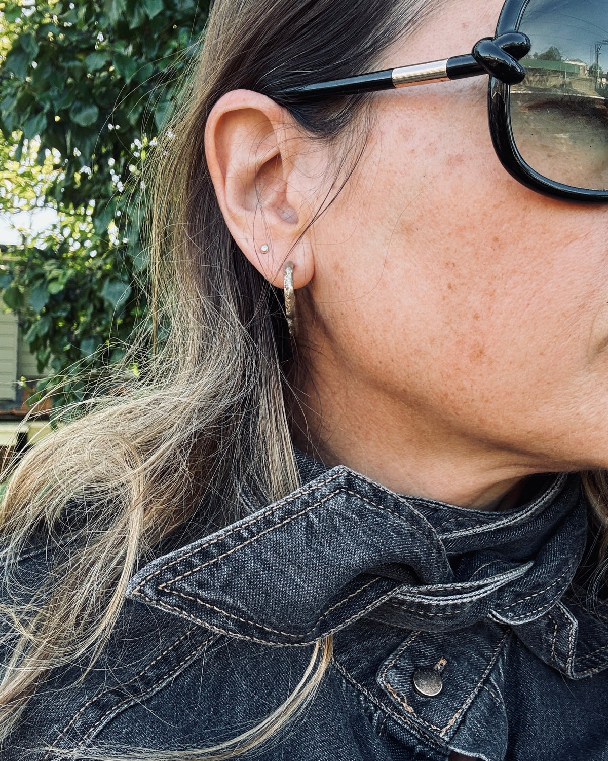 A close-up side profile of a woman wearing a small gold hoop earring, with her ear clearly visible and sunglasses partially covering her face.