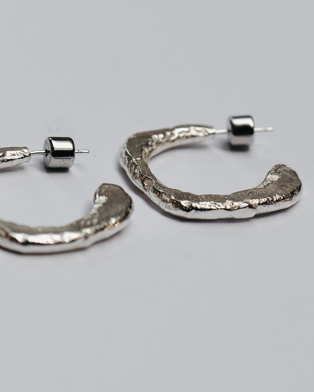 A pair of textured silver hoop earrings with a hammered, organic finish and post-back closures, displayed on a neutral light gray background.