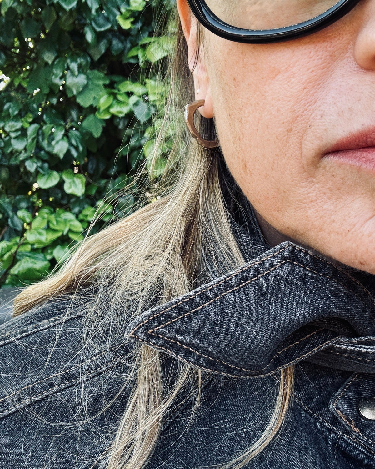 A close-up lifestyle shot of a person wearing a small, rustic brown wooden hoop earring, a black denim jacket, and black-rimmed sunglasses, with green foliage in the background.