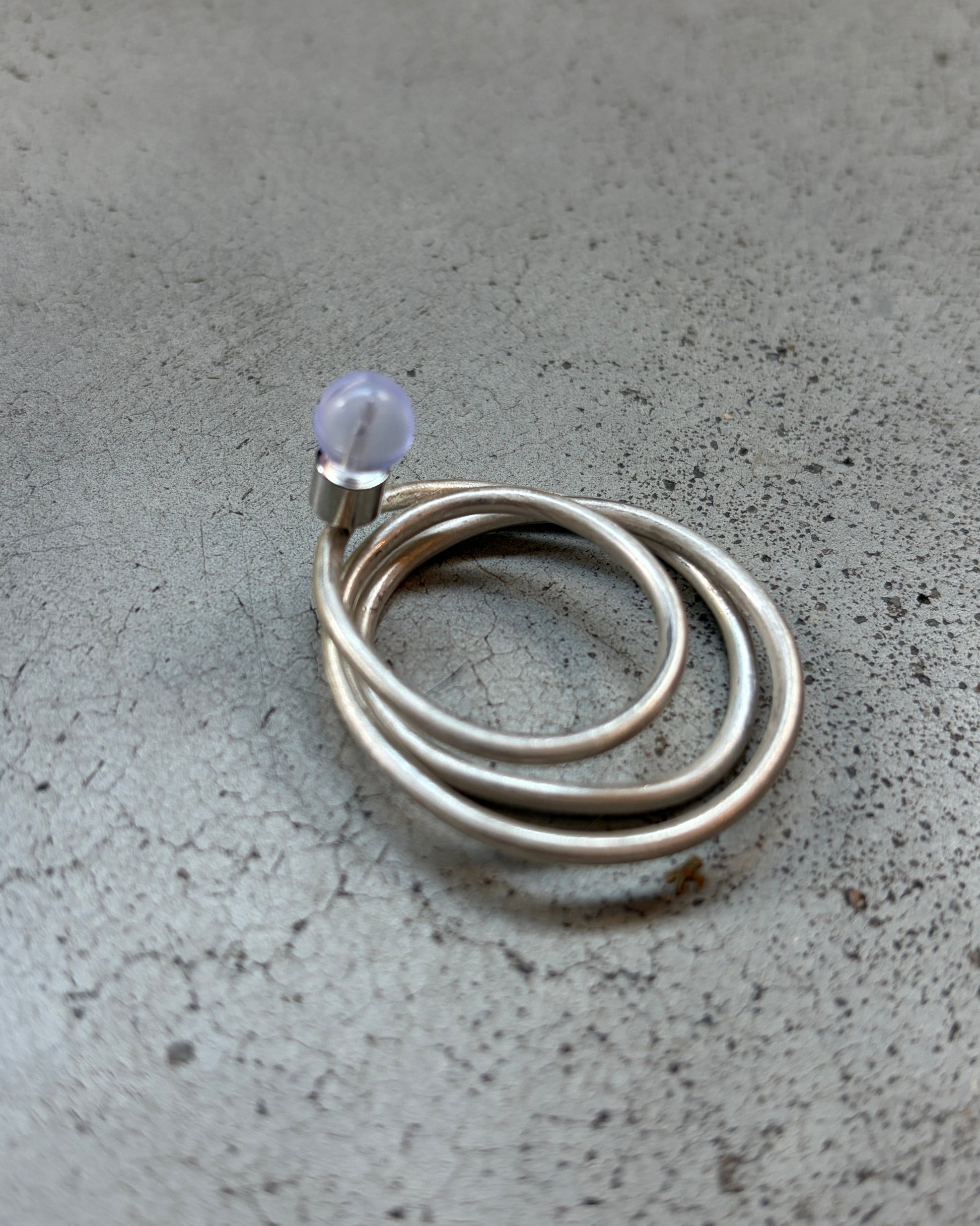 A silver-toned coiled ring with a small clear faceted bead, photographed on a textured gray concrete surface.