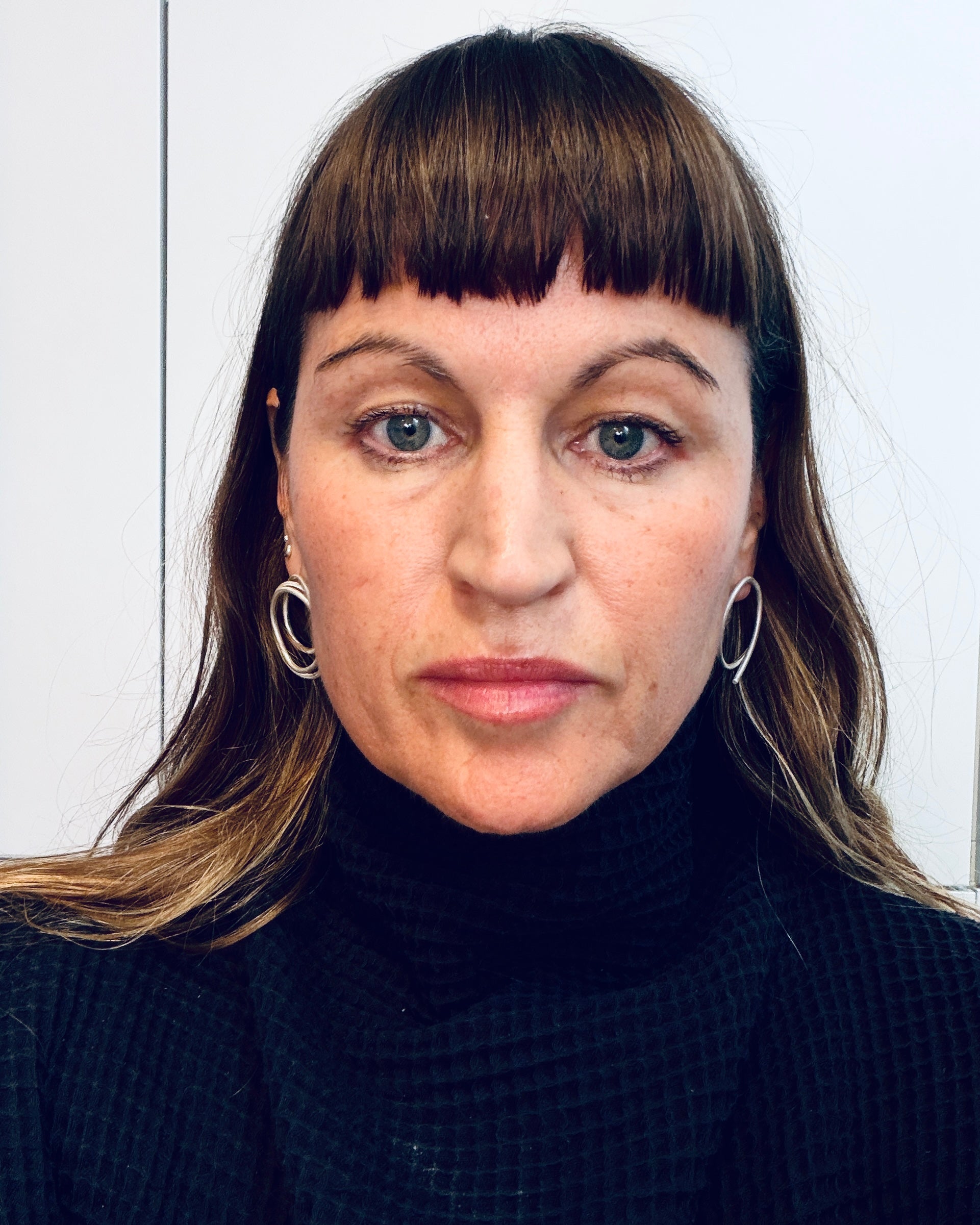 A close-up portrait of a woman with brown hair and straight bangs, wearing silver hoop earrings and a black textured turtleneck sweater, set against a plain light background.