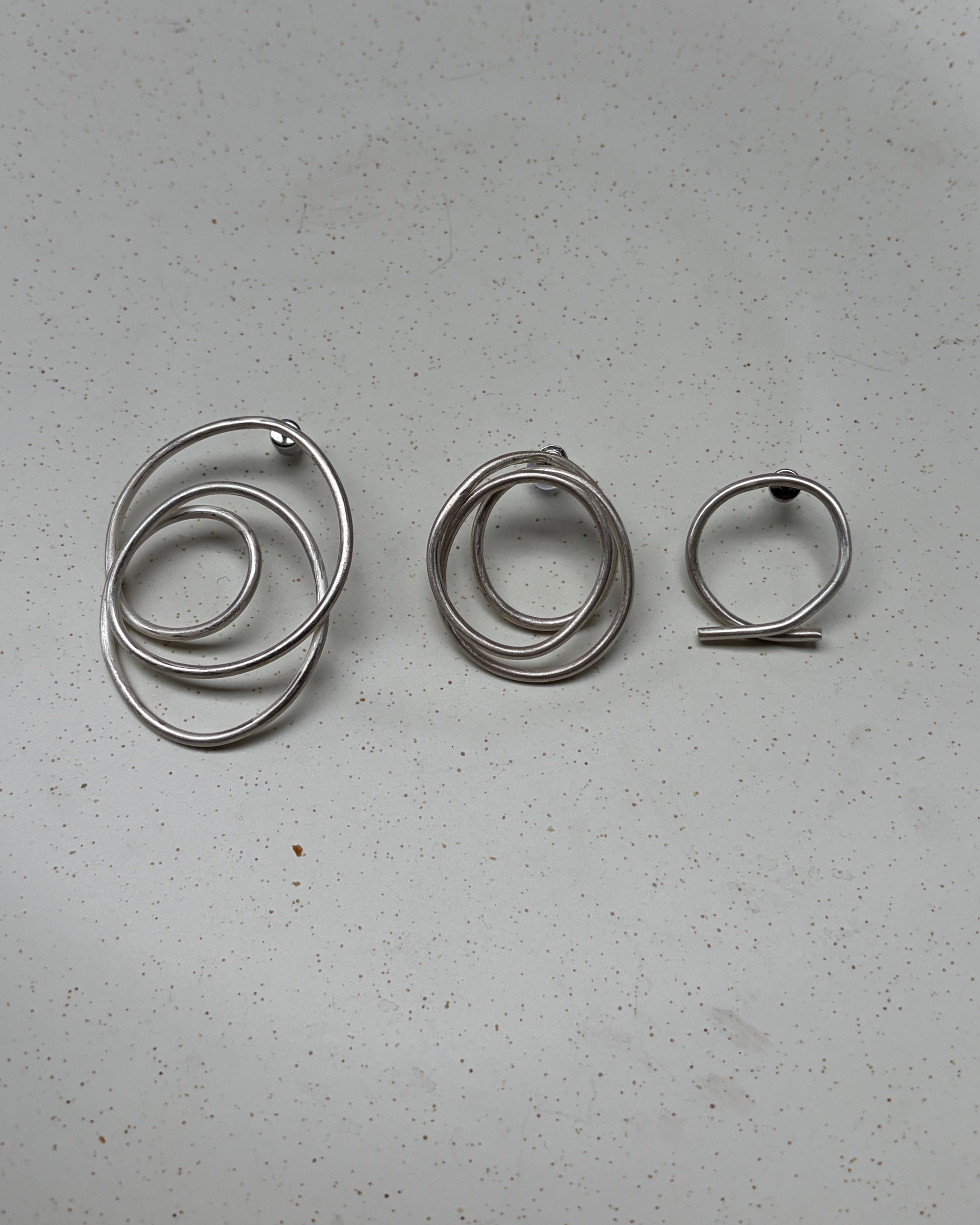 Three silver metal wire hoop earrings arranged vertically on a light gray speckled surface.