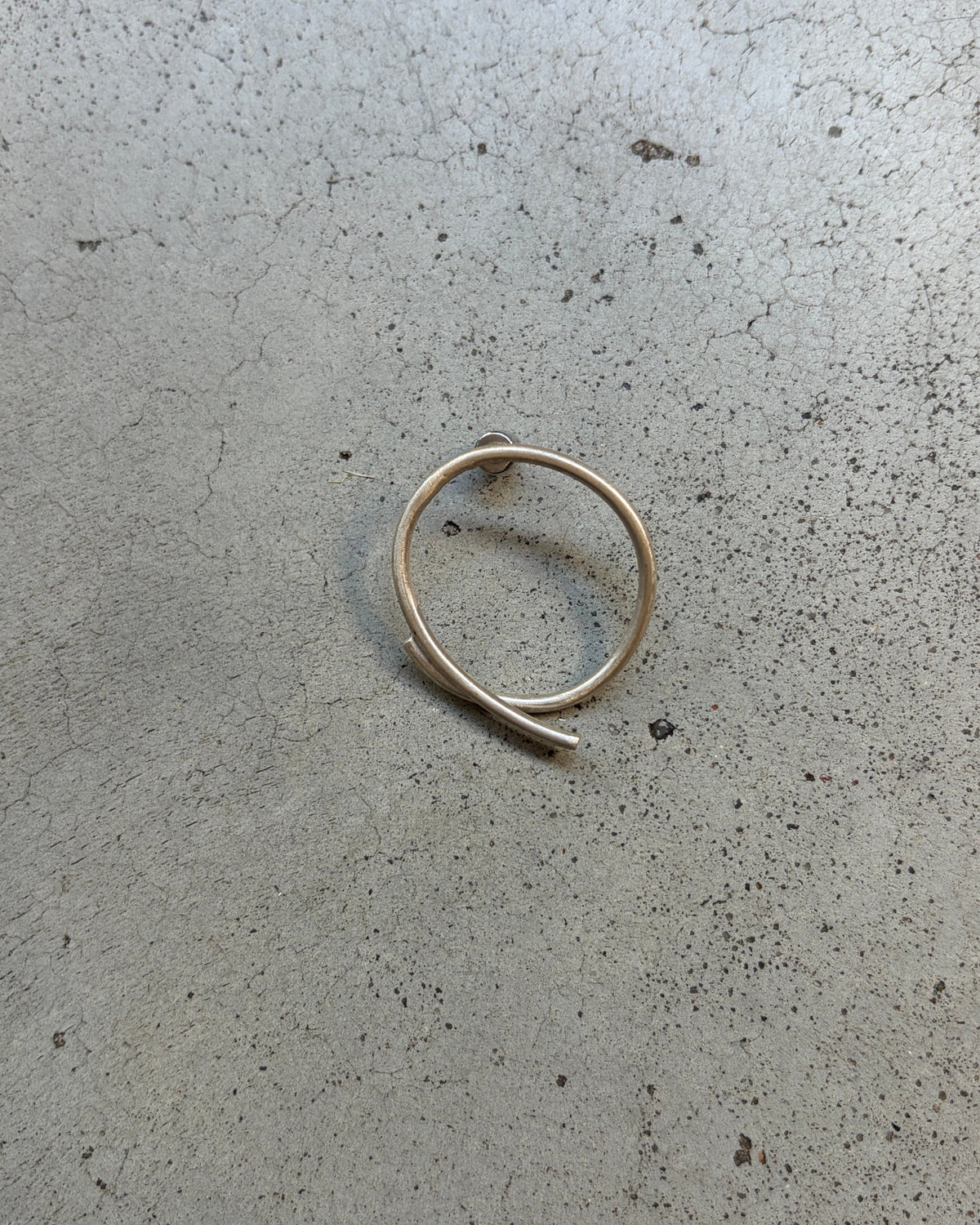 A thin, polished silver ring shaped like a spiral or coiled loop, lying on a textured gray concrete surface.