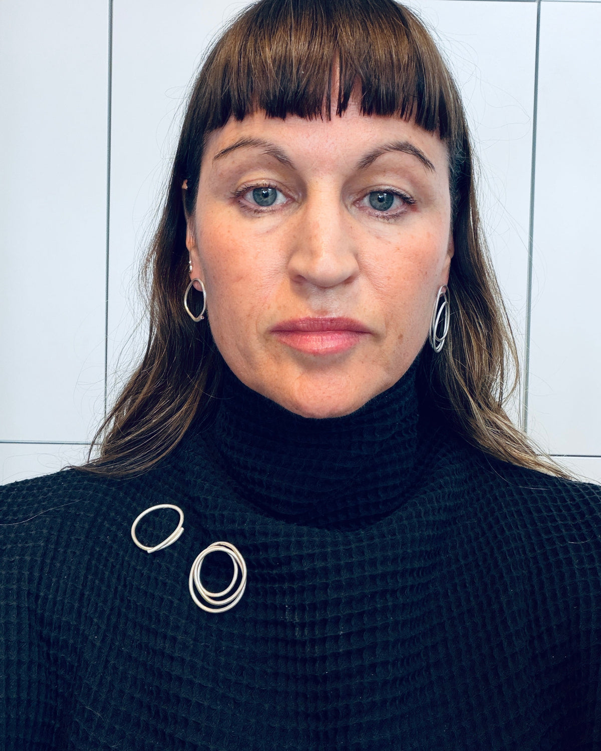 A woman with brown hair and bangs wearing a black waffle-knit turtleneck sweater with silver ring details on the shoulders, set against a white tiled wall.