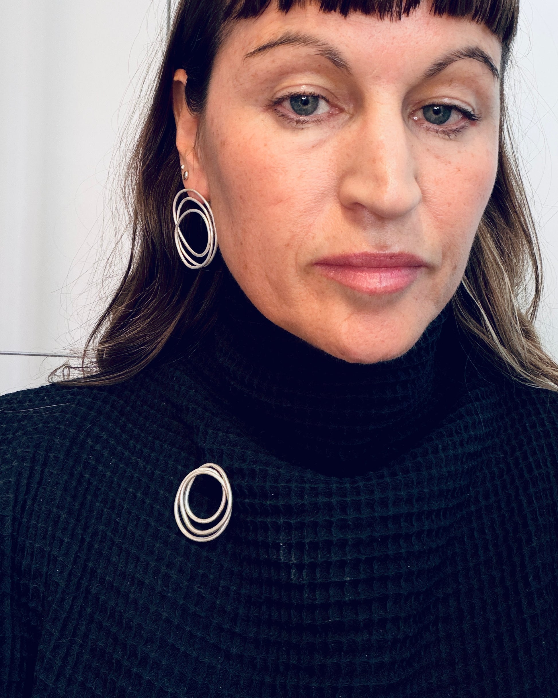 A close-up of a woman wearing silver-toned concentric circle hoop earrings and a matching silver ring detail on her black waffle-knit turtleneck sweater.