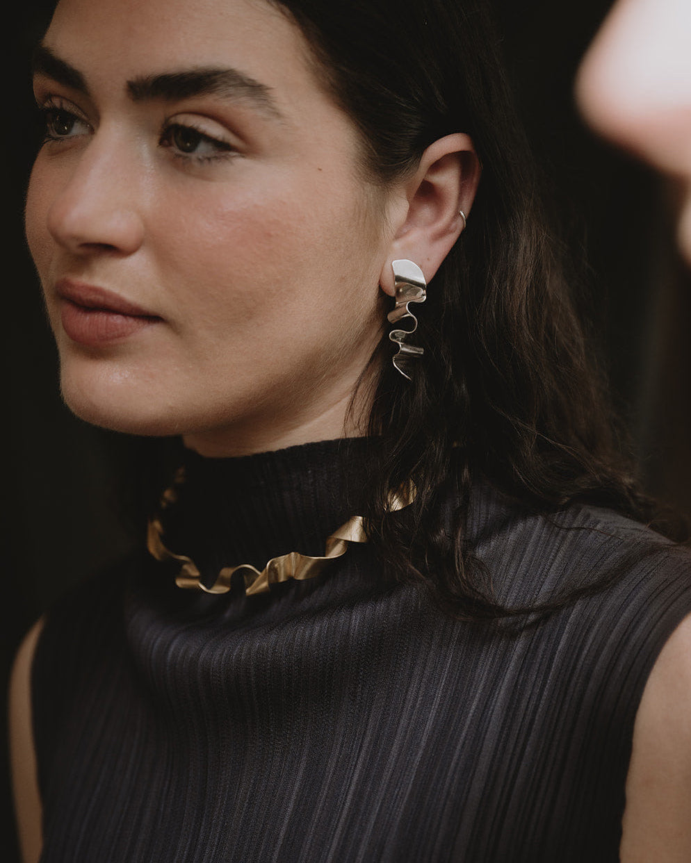 A close-up lifestyle shot of a woman wearing a black pleated mock-neck top and a chunky gold ruffled collar necklace, with a matching gold and white spiral drop earring visible.