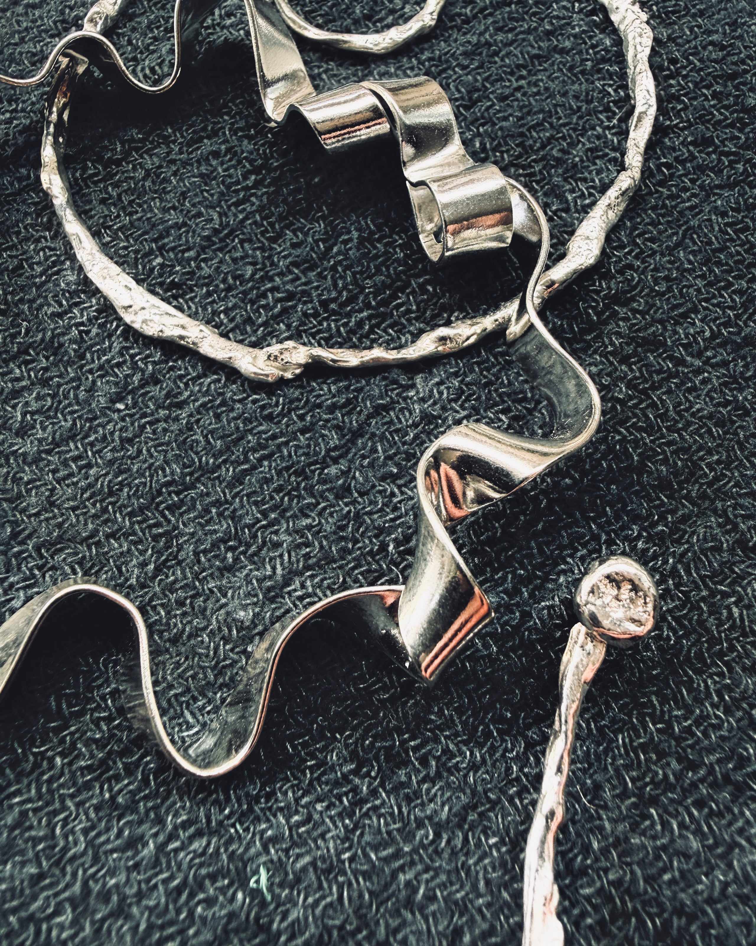 A close-up of a silver-toned jewelry piece with sculptural, wavy ribbon-like elements and a textured organic border, laid on a dark woven fabric background.