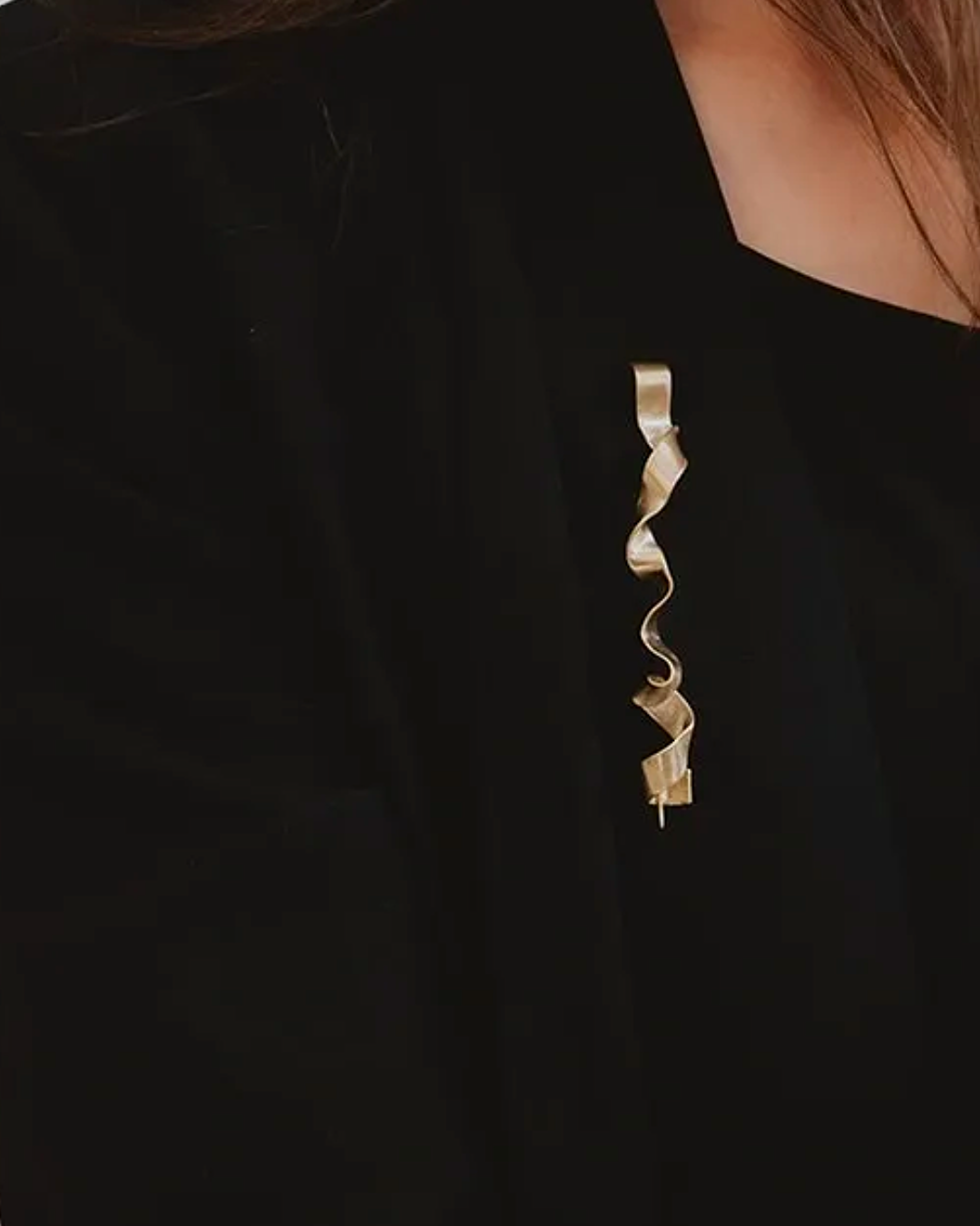 A close-up of a person wearing a black top and a long, wavy, ribbon-like gold brooch pinned to the chest.