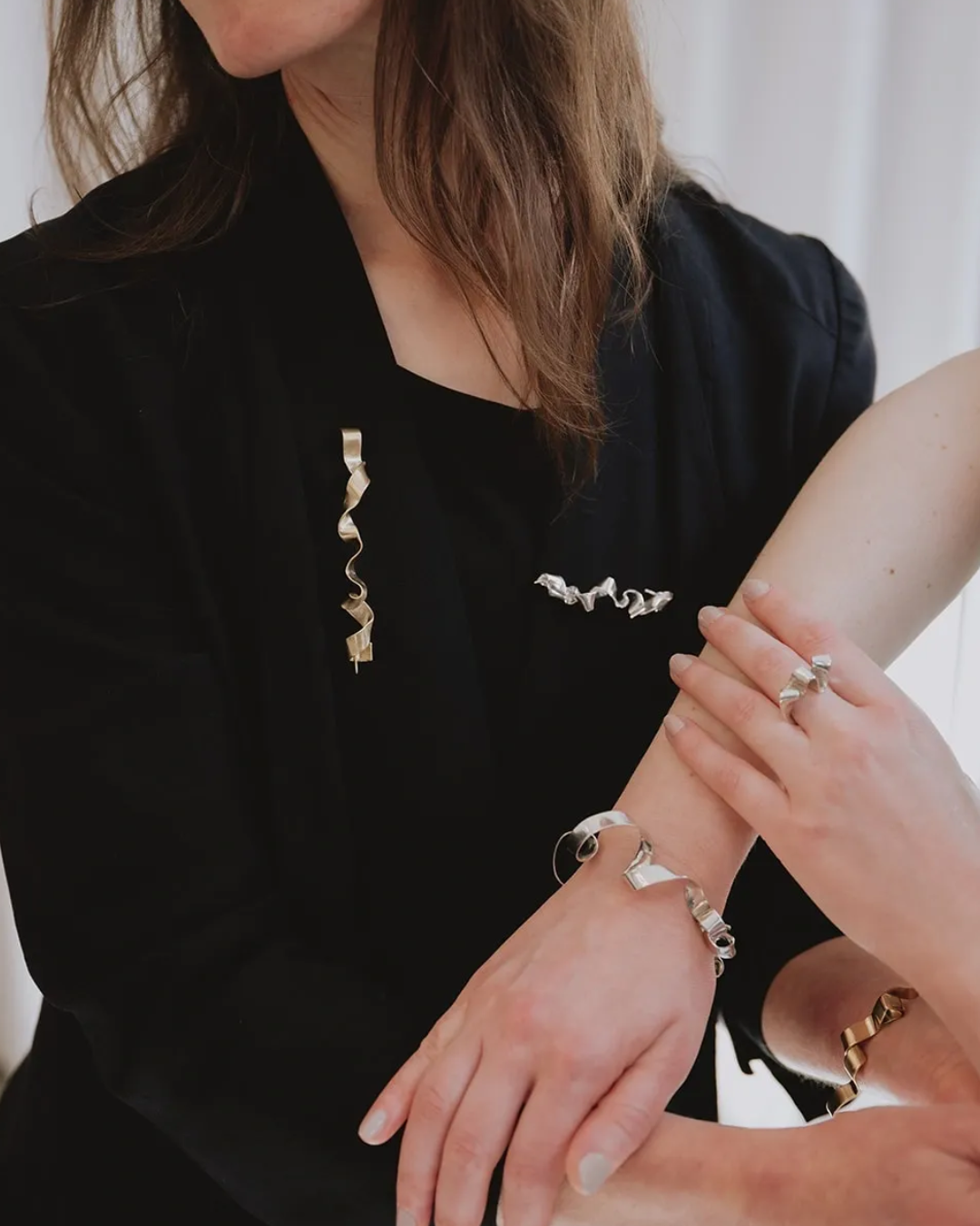 A close-up lifestyle shot of a model wearing sculptural silver and gold jewelry, including a long ribbon-shaped brooch, matching bracelets, and rings, styled with a black velvet blazer.