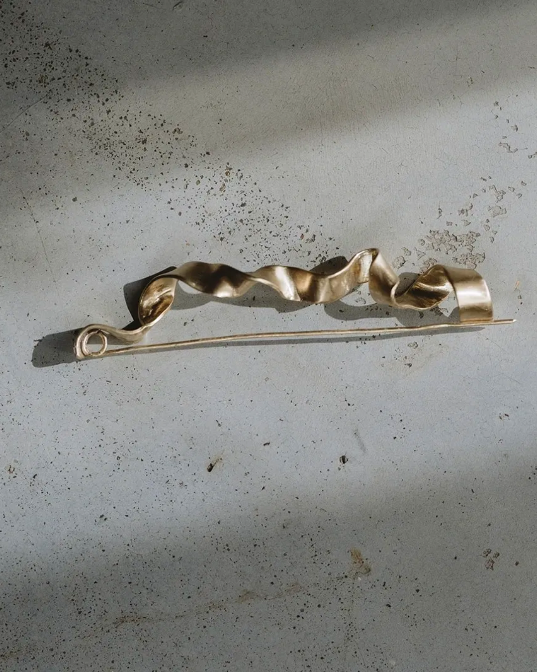 A gold-toned metal pin with a wavy, ribbon-like top section and a straight pin body, resting on a textured gray concrete surface with soft shadows.