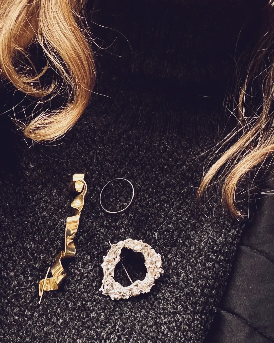 Three jewelry items arranged on a dark textured surface: a gold-toned curled ribbon hair accessory, a thin silver hoop earring, and a textured cream-colored floral hair pin. Blonde hair is visible at the top and right edges.