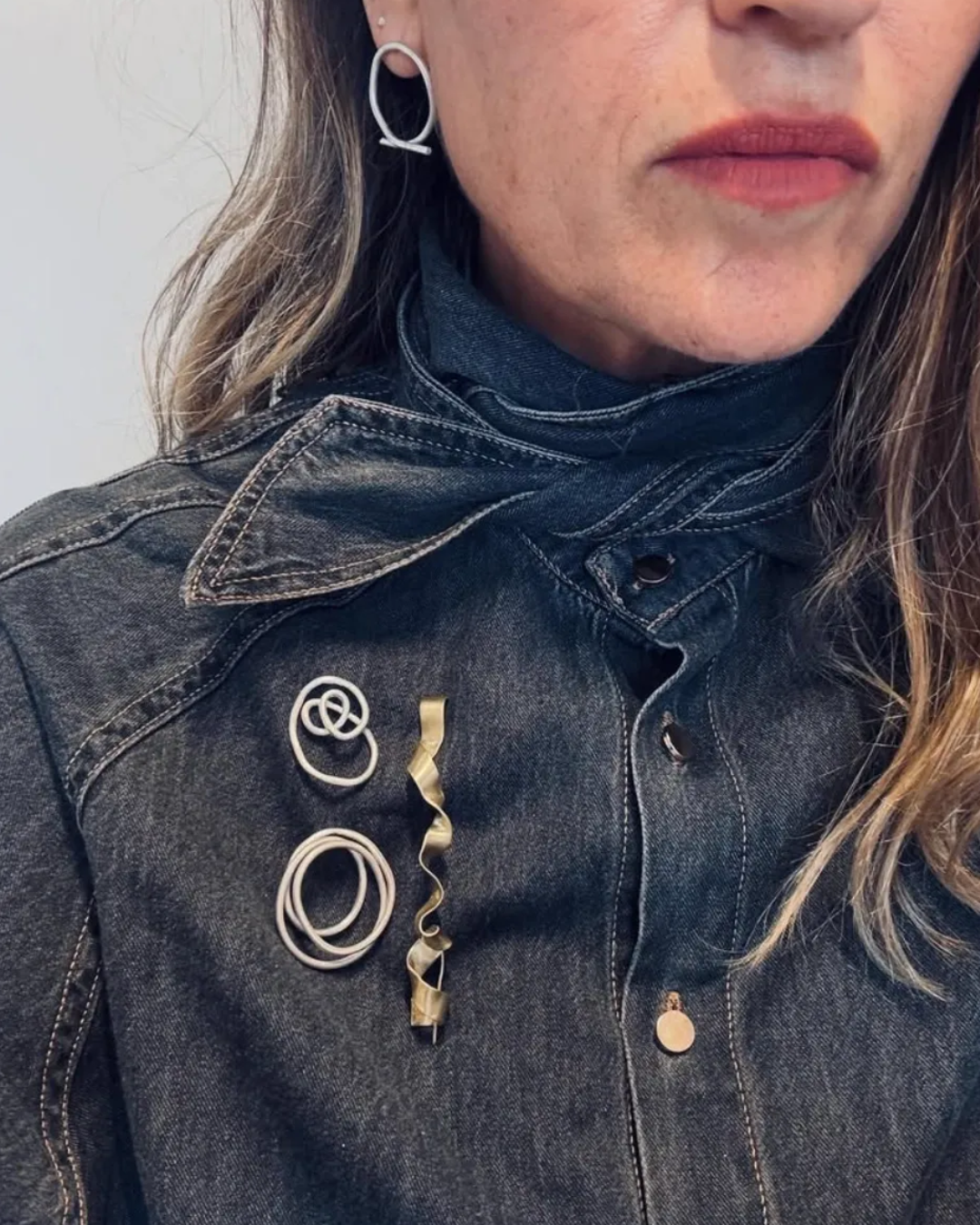 A close-up of a person wearing a dark denim jacket with several gold and silver metal hair pins and barrettes pinned to the chest, along with a silver hoop earring.