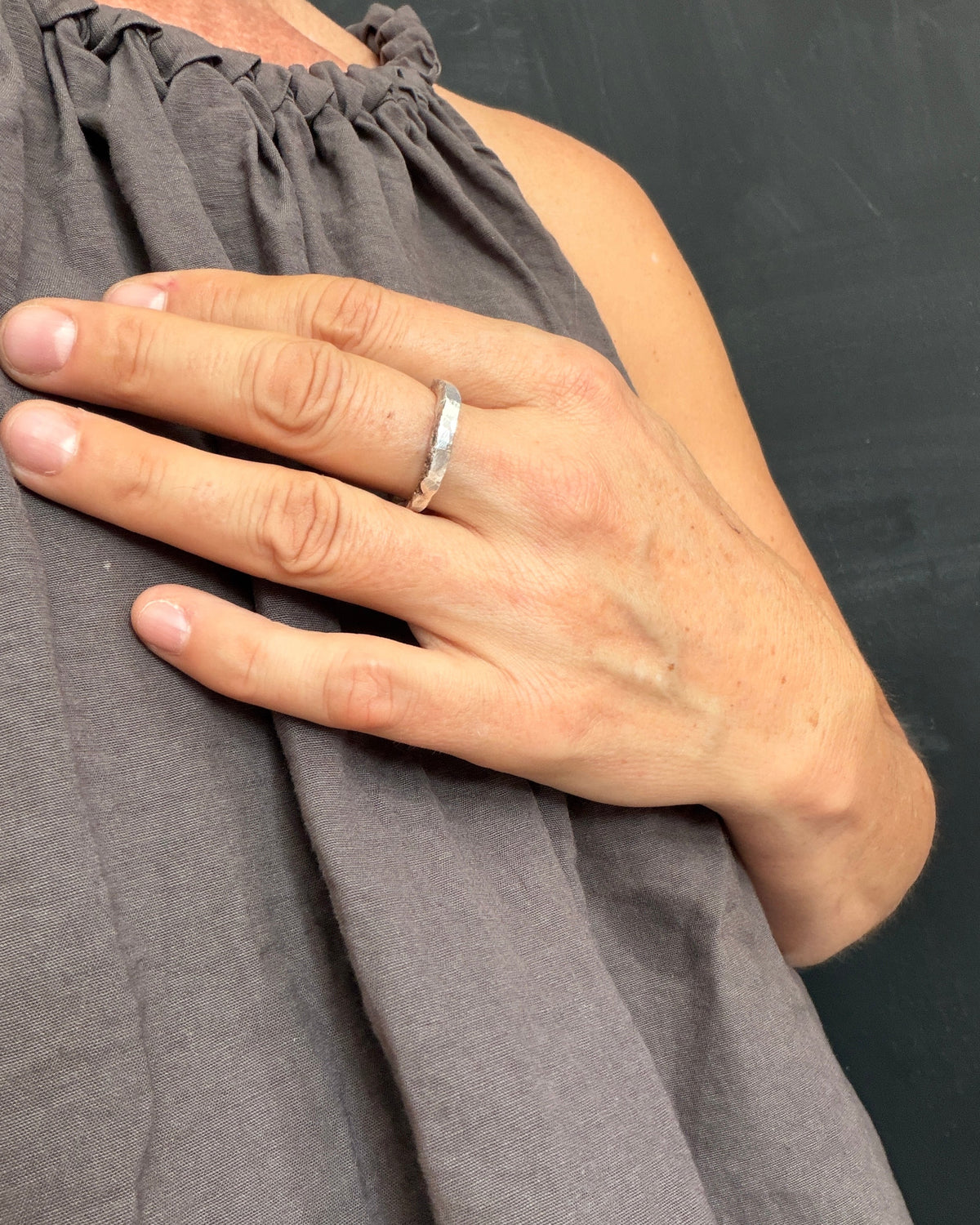 A close-up of a person's hand wearing a slim silver band ring, resting on their chest against a dark gray background.