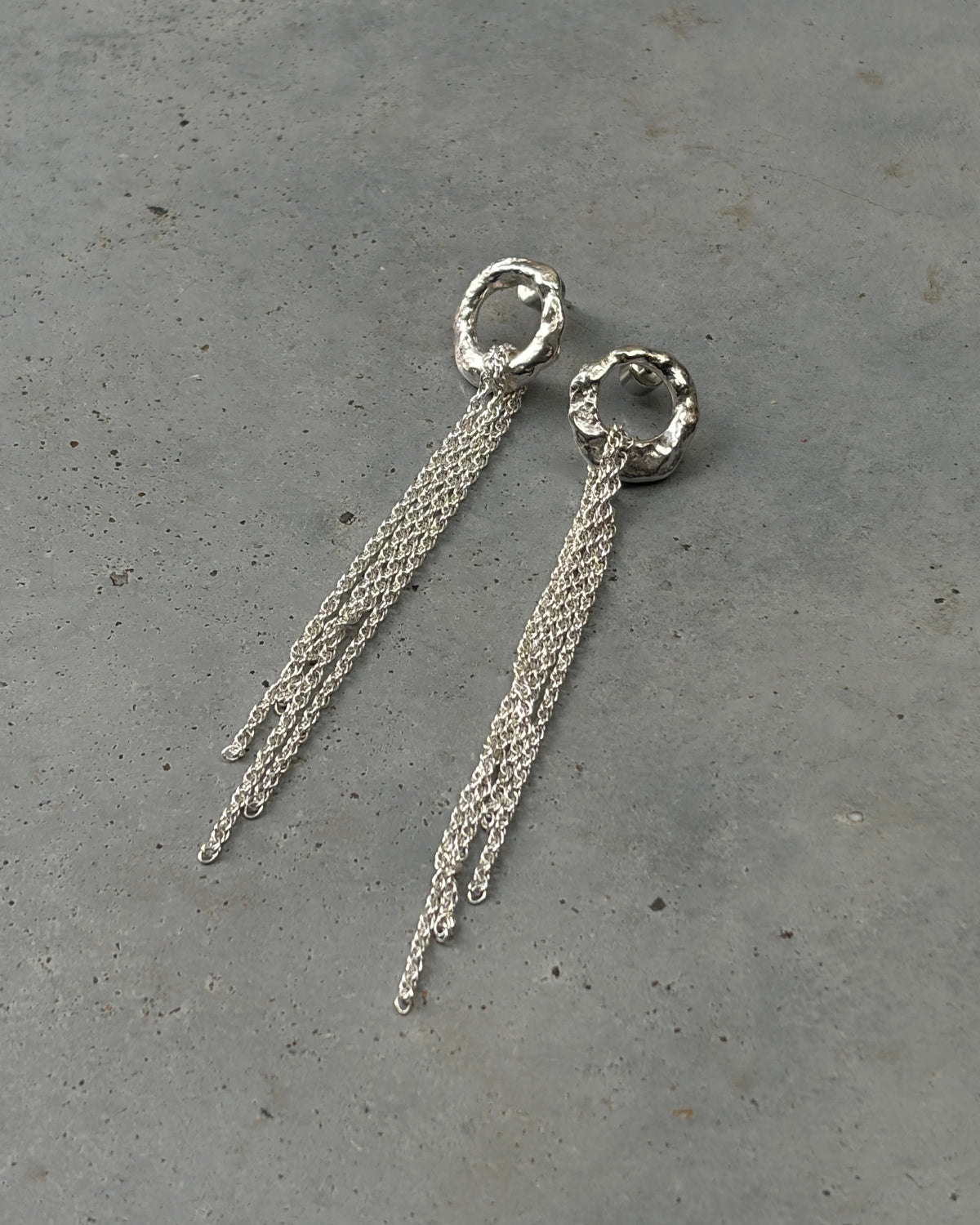 A pair of silver dangle earrings with textured circular hoops and three fine chain drops, displayed on a gray concrete surface.