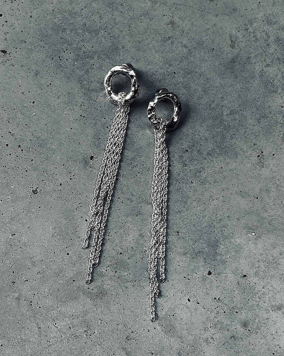 close up of charcoal cast silver hoop earrings with a silver rope chain hanging from them sitting on a concrete floor