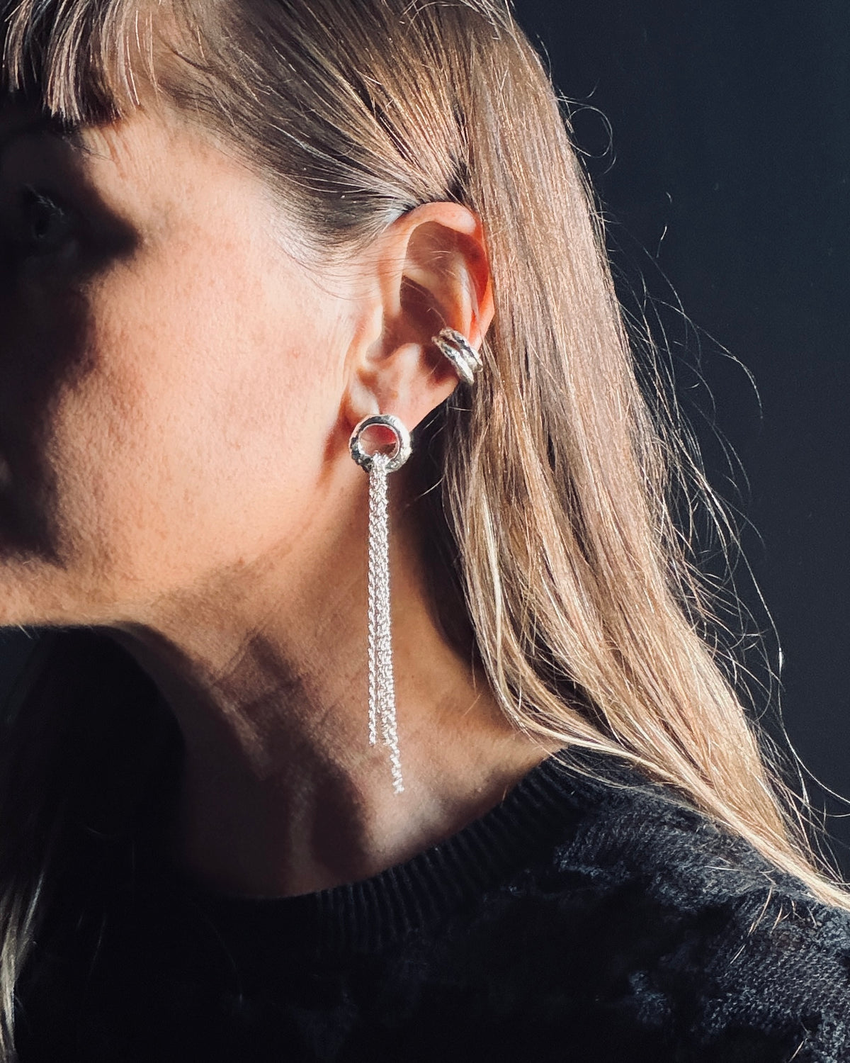 A close-up side profile of a woman wearing a silver hoop earring with a dangling chain and a matching silver ear cuff against a dark background.