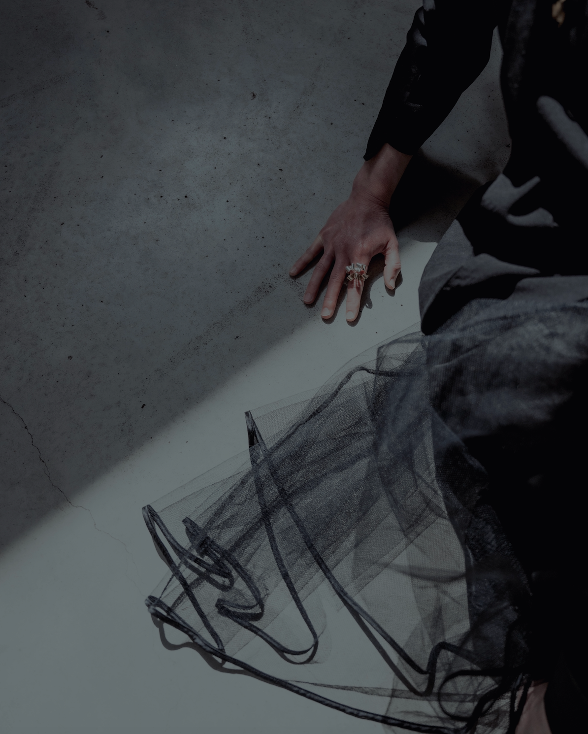 A close-up, high-angle shot of a person's hand resting on a concrete floor next to a flowing black tulle skirt or dress.