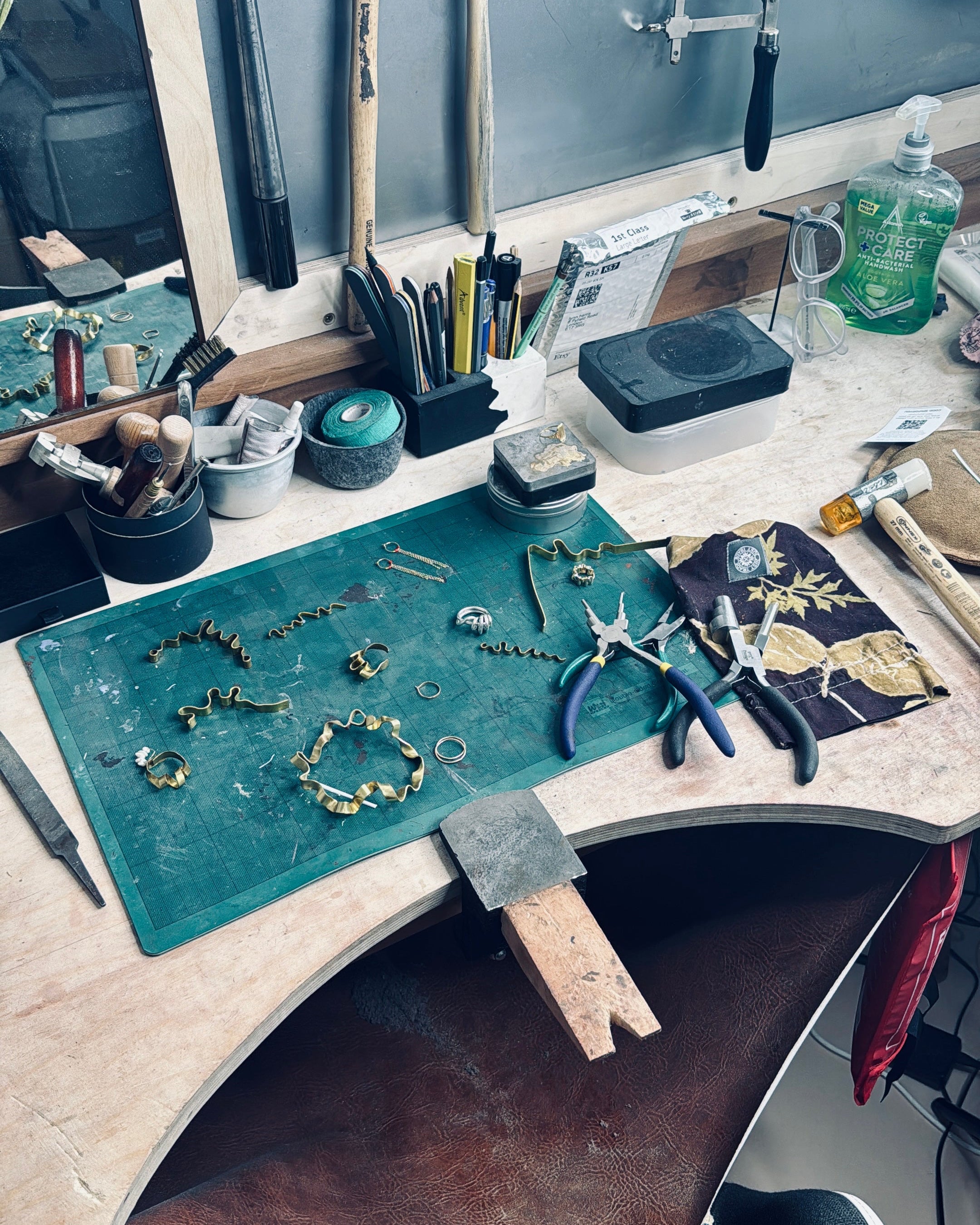 A detailed view of a jewelry maker's workbench featuring a green soldering mat with jewelry components, pliers, a hammer, and various tools on a stone work surface.