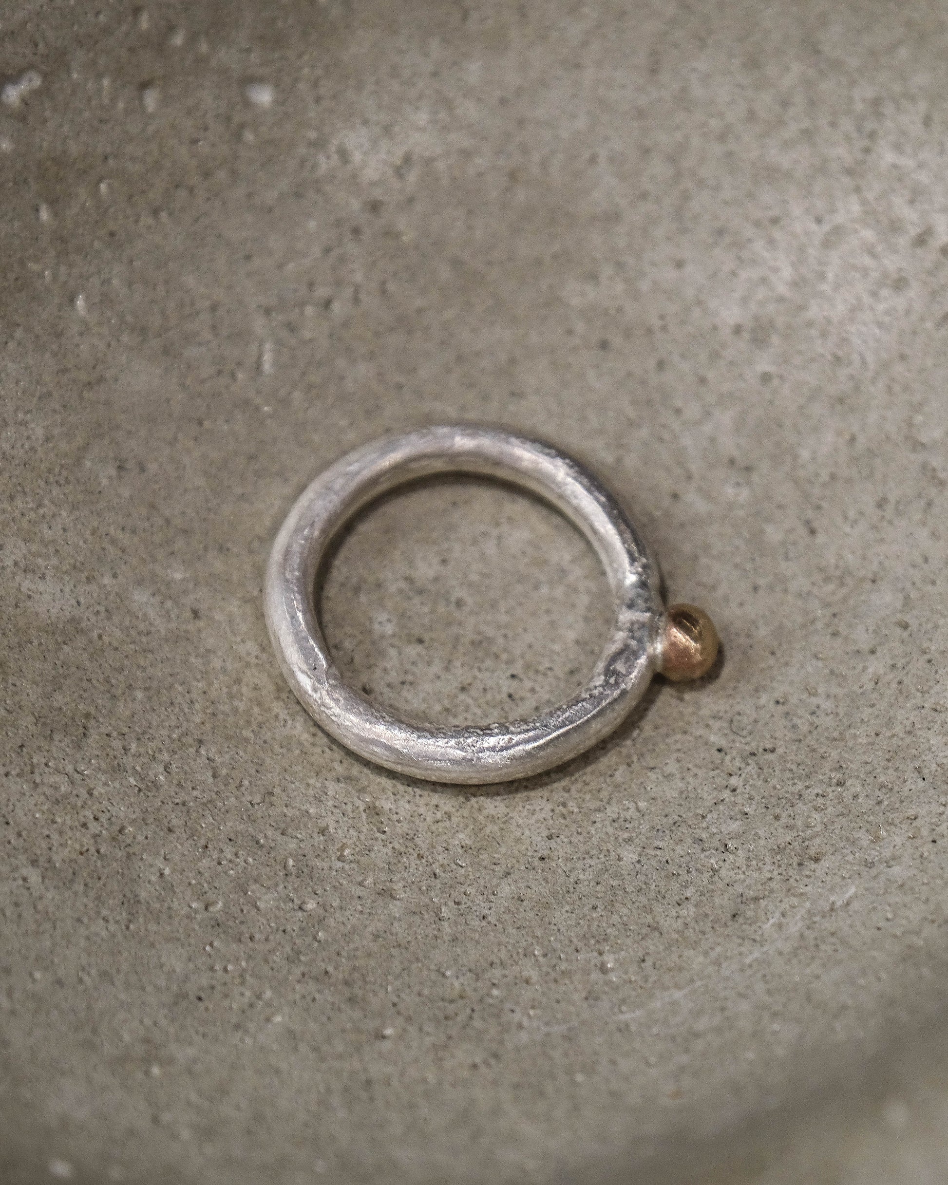 A silver-toned metal ring with a textured, hammered finish and a small brass-colored loop at the top, resting inside a textured gray concrete bowl.