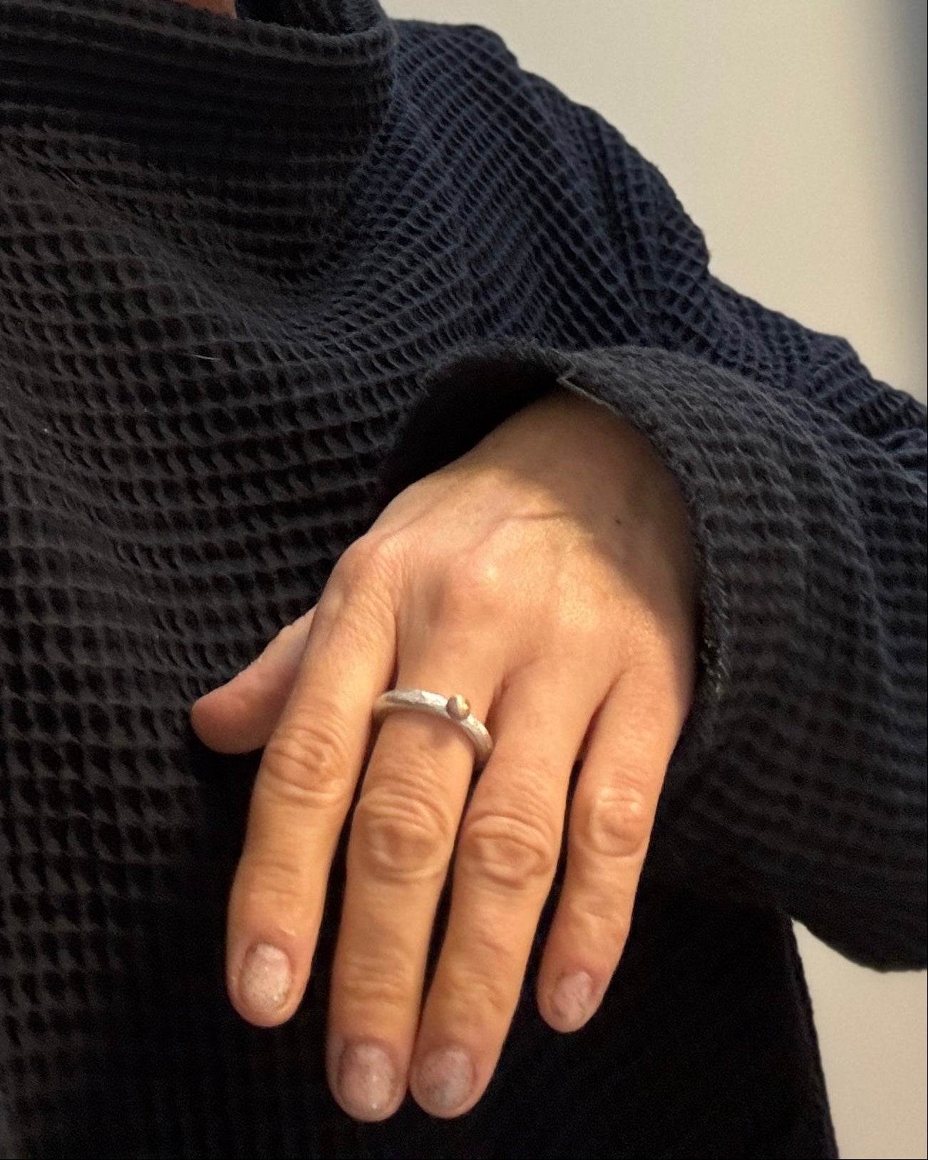 A close-up of a person's hand wearing a simple silver band ring, shown against a dark textured waffle-knit sweater.