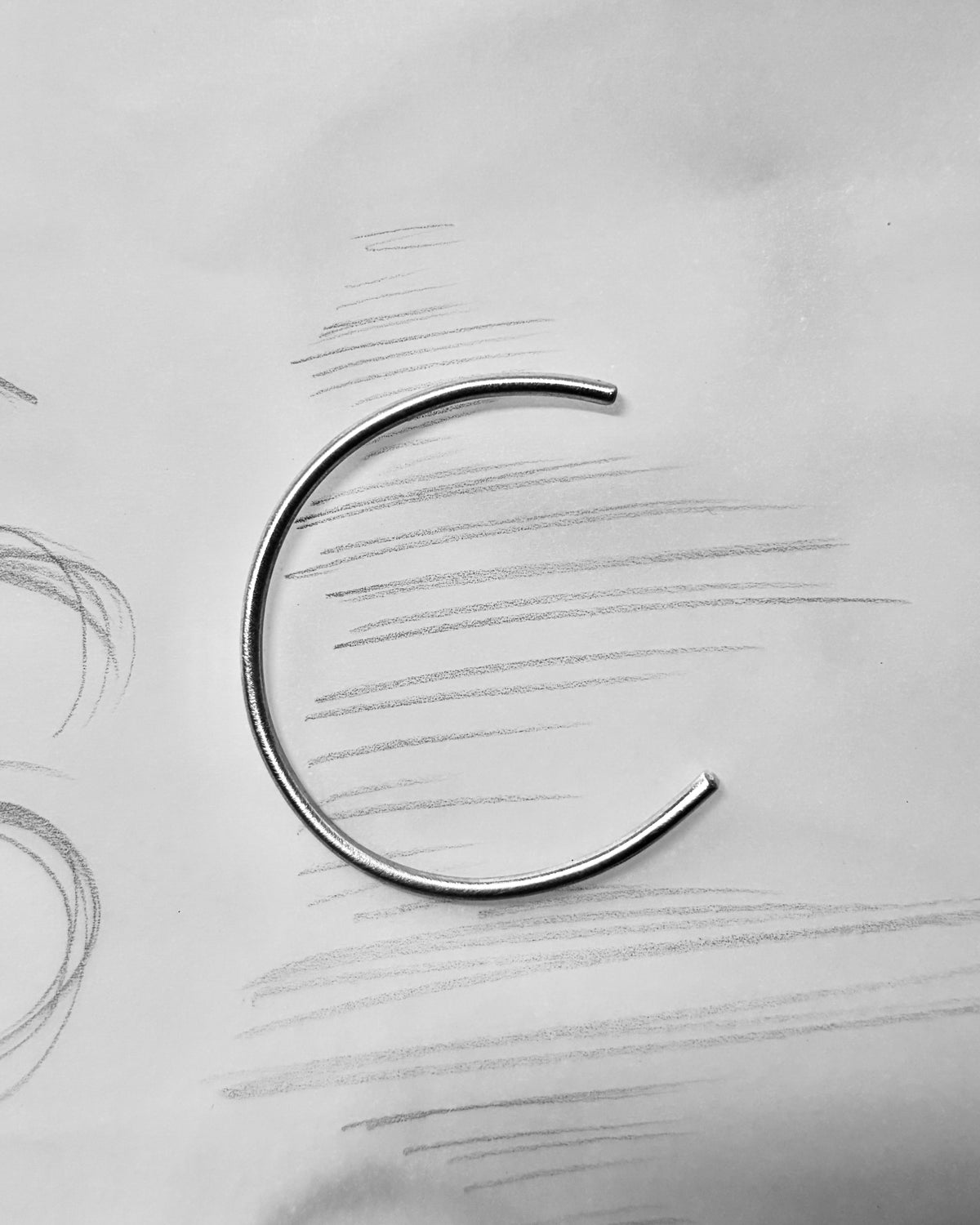 A single thin metal C-shaped ring or loop with open ends, photographed on a light gray paper-like surface with faint pencil sketch lines in the background.