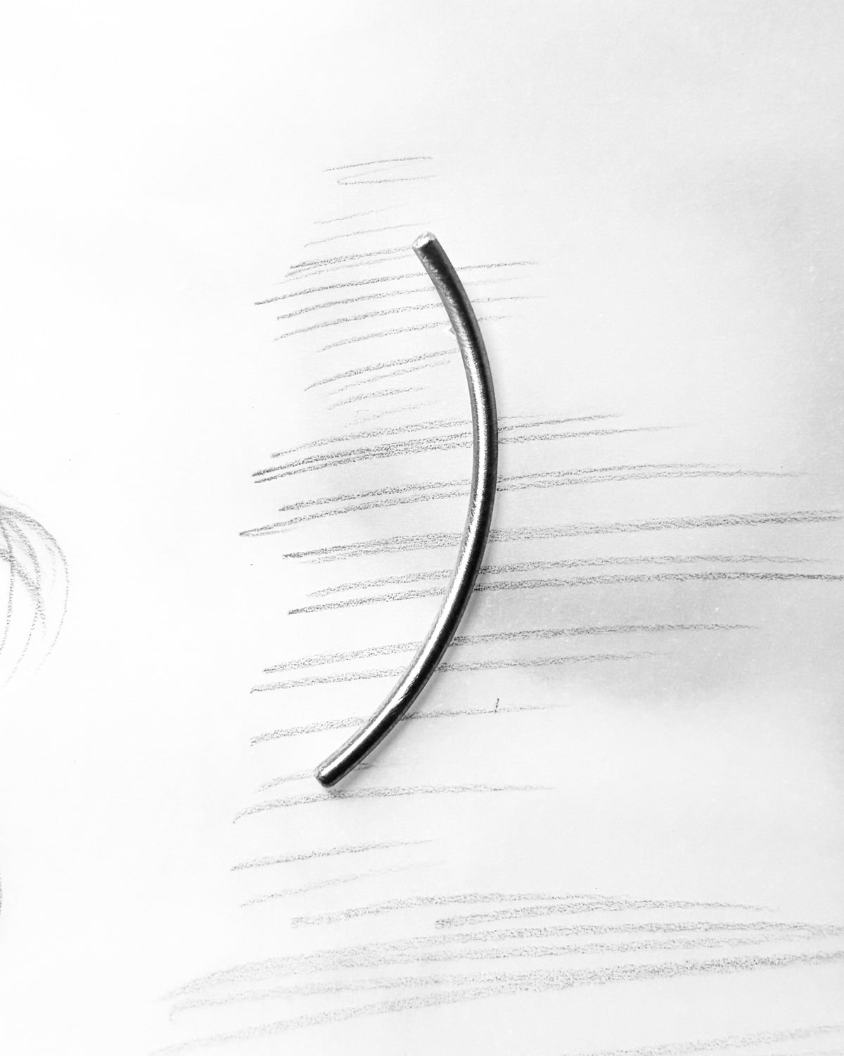 A single curved metallic bar, appearing silver-toned, placed on a white paper background with faint pencil sketch lines.