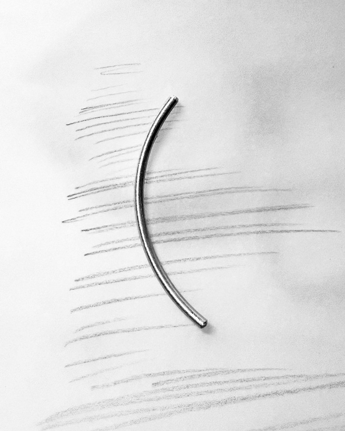 A single curved metallic bar, likely a jewelry finding or hardware piece, placed on a white background with faint pencil sketch lines.